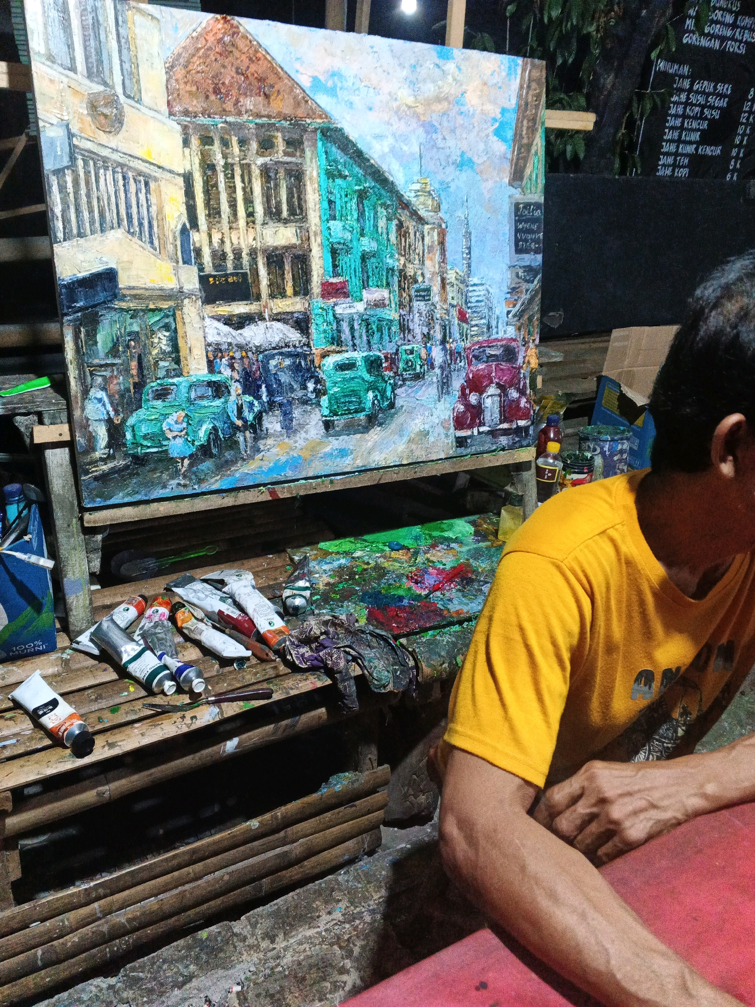 The image depicts an artist in their studio, situated outdoors, likely in Ungaran, Indonesia. The artist, wearing a yellow t-shirt, is positioned in the foreground, with their right arm resting on a red surface. Their focus appears to be on a large, textured oil painting displayed on an easel. The painting itself is a vibrant depiction of a street scene, featuring colorful buildings, vintage cars, and figures in a bustling urban environment.
Scattered around the easel are various art supplies, including tubes of paint, brushes, and what appears to be a palette knife, indicating the ongoing nature of the artistic process. The background reveals a dark, natural setting, possibly a garden or wooded area, with a blackboard displaying a menu of food and drinks, suggesting the studio might be part of an outdoor cafe or a community art space. The overall impression is one of a creative environment, capturing a moment of artistic contemplation and work.