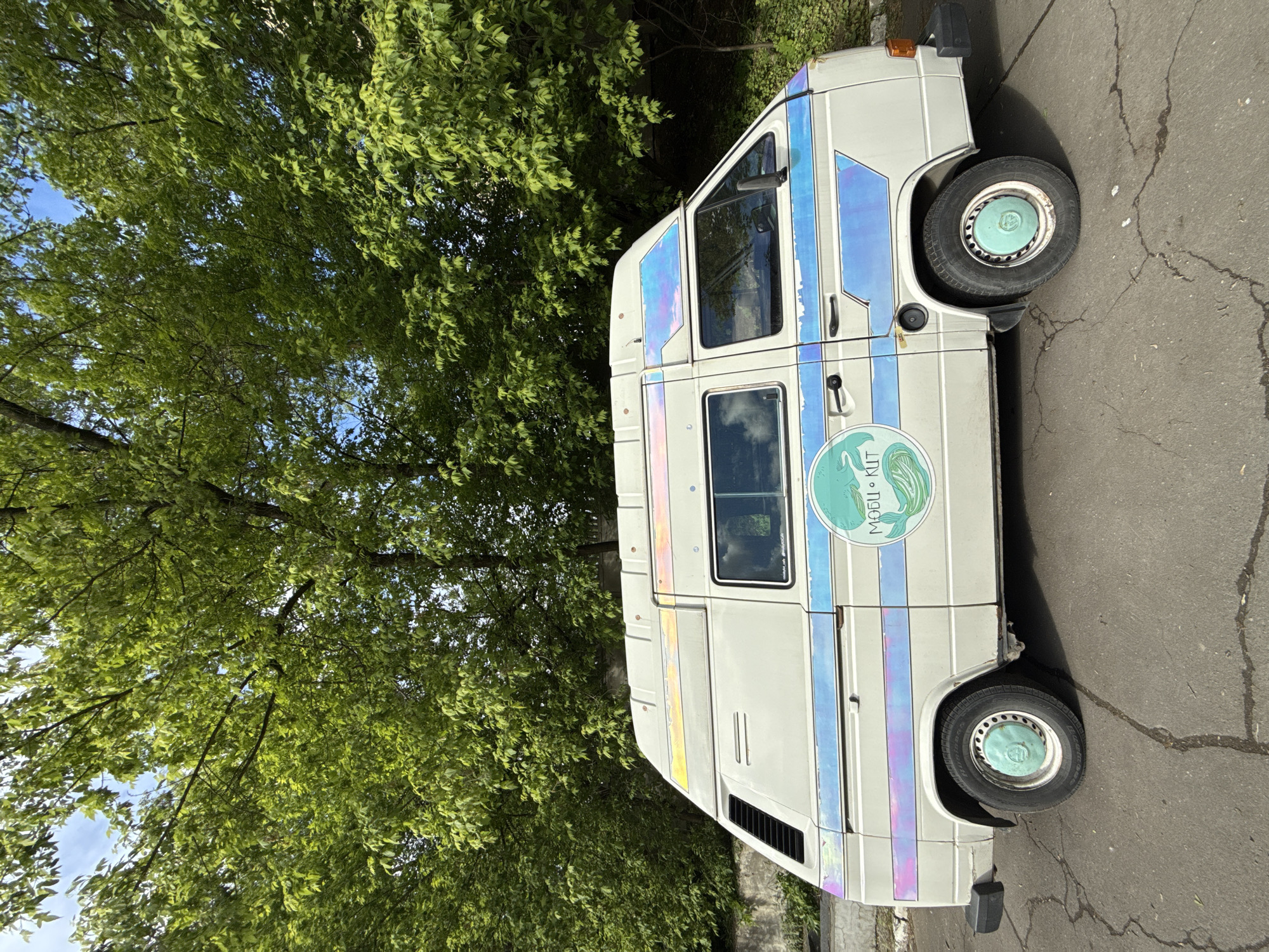 An eye-level shot presents the side of a white van parked on asphalt pavement near a tree. The van features iridescent stripes and a circular logo with Cyrillic text and an image of a whale. The tires have light blue hubcaps. The background includes foliage, suggesting a park or a tree-lined street. The location is indicated as Moscow, Russia, based on the Cyrillic text and the environment depicted.