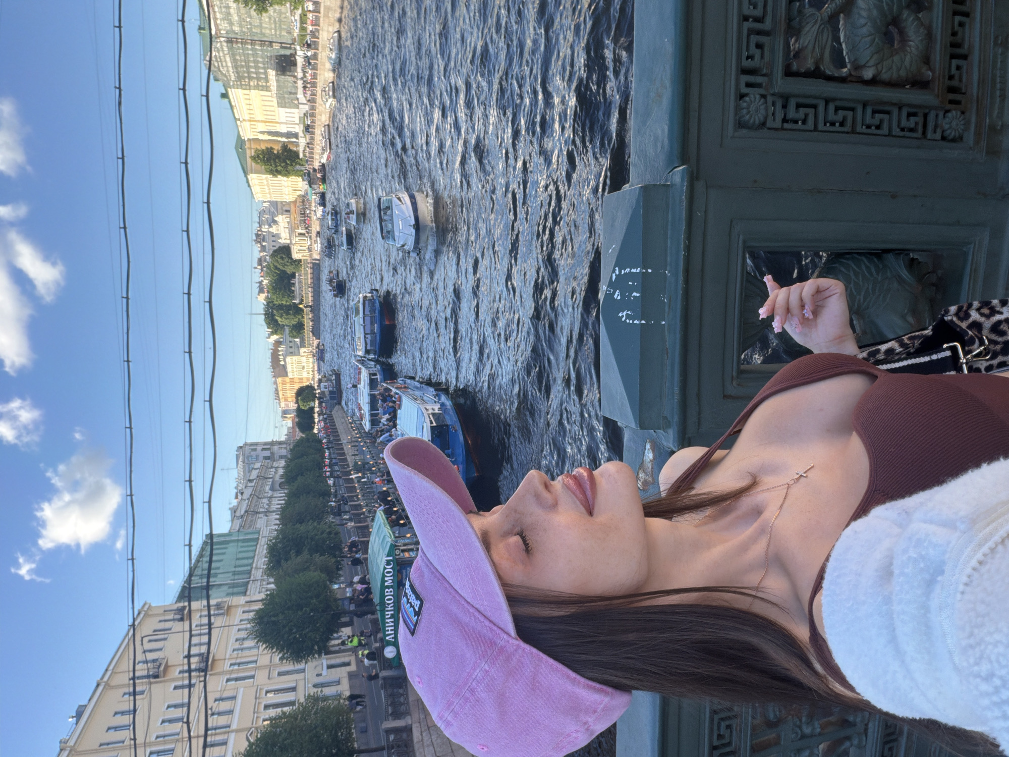 The image shows a young woman with long dark hair, wearing a pink baseball cap and a brown top with a white fuzzy jacket draped over her shoulders. She is standing on a bridge in Saint Petersburg, Russia, with a canal and several boats in the background. Buildings line the canal, and the sky is blue with some clouds. The woman is looking to her left, with her eyes closed and a slight smile on her face. She is holding a cigarette in her right hand. The bridge has a decorative green railing. The scene appears to be a sunny day.