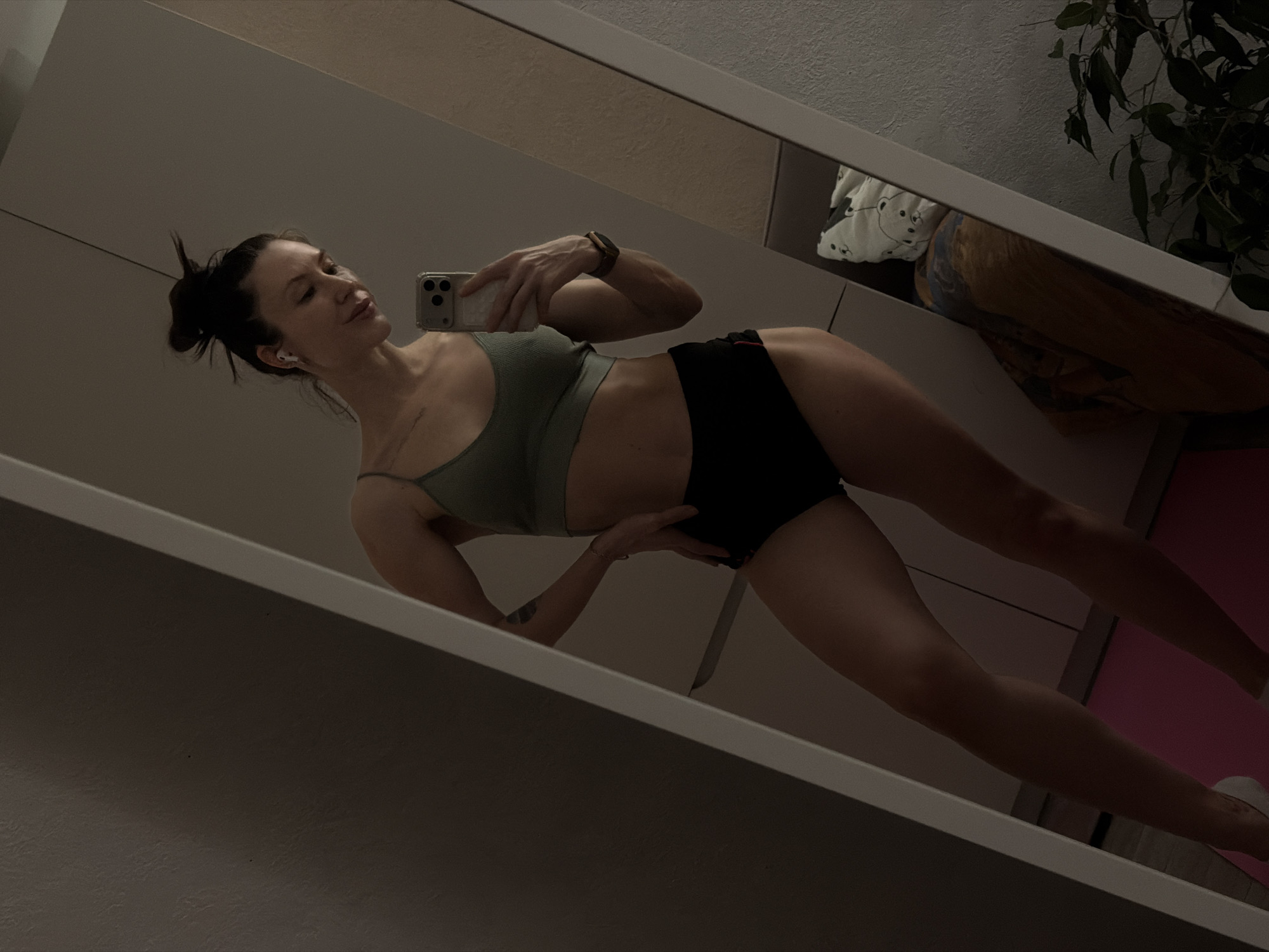 A medium shot depicts a female subject in a mirror selfie. She has her hair pulled back in a messy bun and wears a light green sports bra and dark shorts. She's holding a smartphone in her right hand, capturing her reflection. Her left hand rests on her hip. The setting includes a mirror, parts of a cabinet, a plant, and a pink yoga mat. Location data for the city of Perm, Russia, is unavailable.
