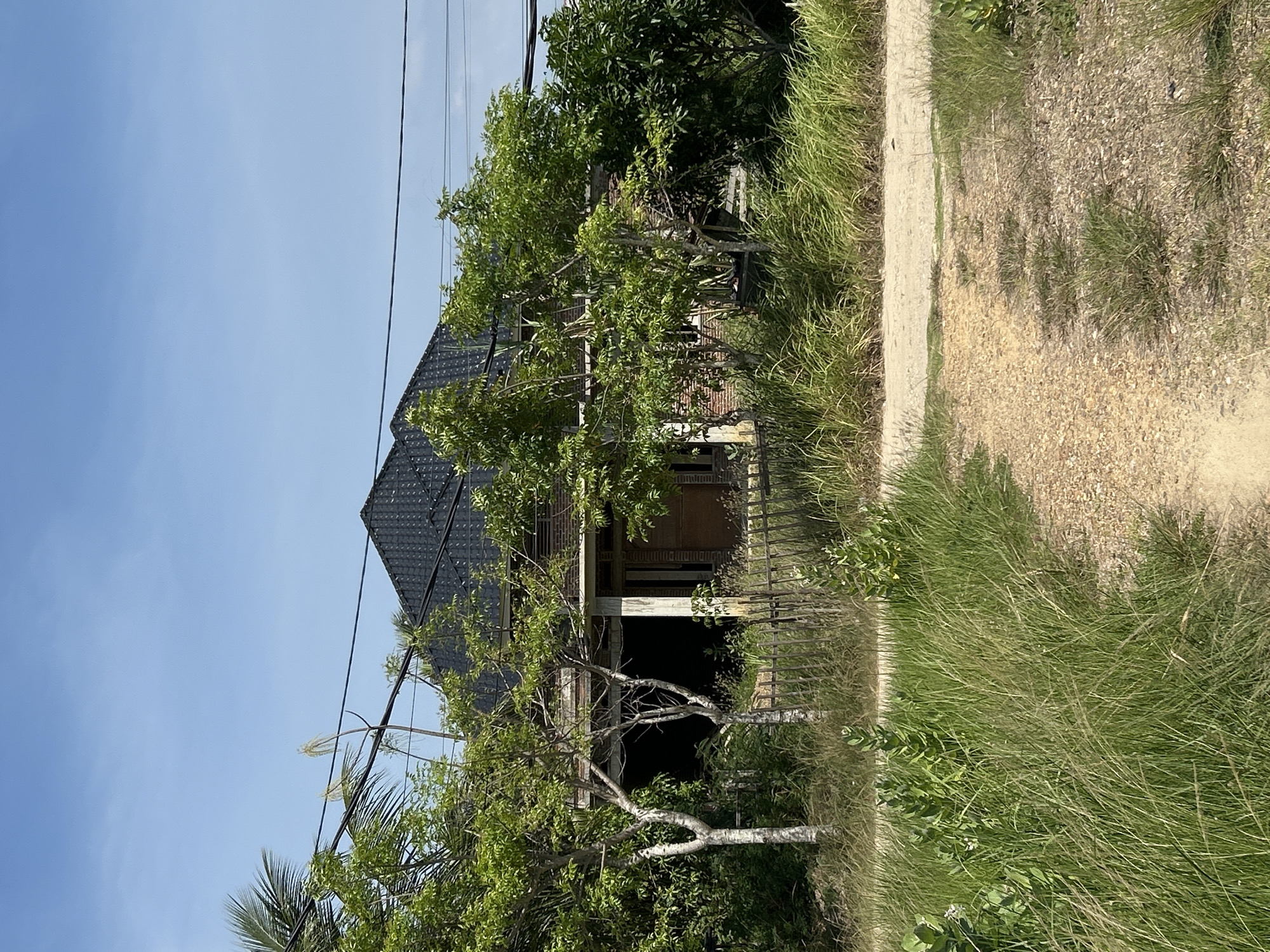 The image depicts an abandoned house in Lhokseumawe, Indonesia. The house has a dark roof and a dilapidated porch with supporting columns. Overgrown vegetation surrounds the house, including tall grasses and trees obscuring the facade. A narrow dirt road runs alongside the property. Overhead, power lines traverse the scene against a clear blue sky. The house appears neglected and possibly vacant. No people are visible.