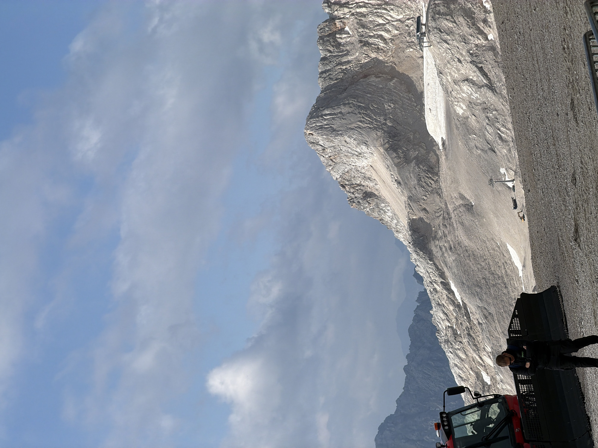 In Garmisch-Partenkirchen, Germany, on a rocky, mountainous terrain, a man dressed in black stands with his arms crossed, facing away from a red snow grooming machine. The machine has a large blade at the front and is parked on a gravelly surface. In the background, a majestic, rugged mountain peak rises, partially covered in snow. The sky above is a mix of blue and gray clouds. Further in the distance, other mountain ranges are visible, some appearing hazy. Two ski lifts are present on the mountain, and smaller construction vehicles are also spotted on a lower part of the slope. The overall scene depicts a high-altitude environment, possibly a ski resort or mountain area, with ongoing maintenance or operational activities.