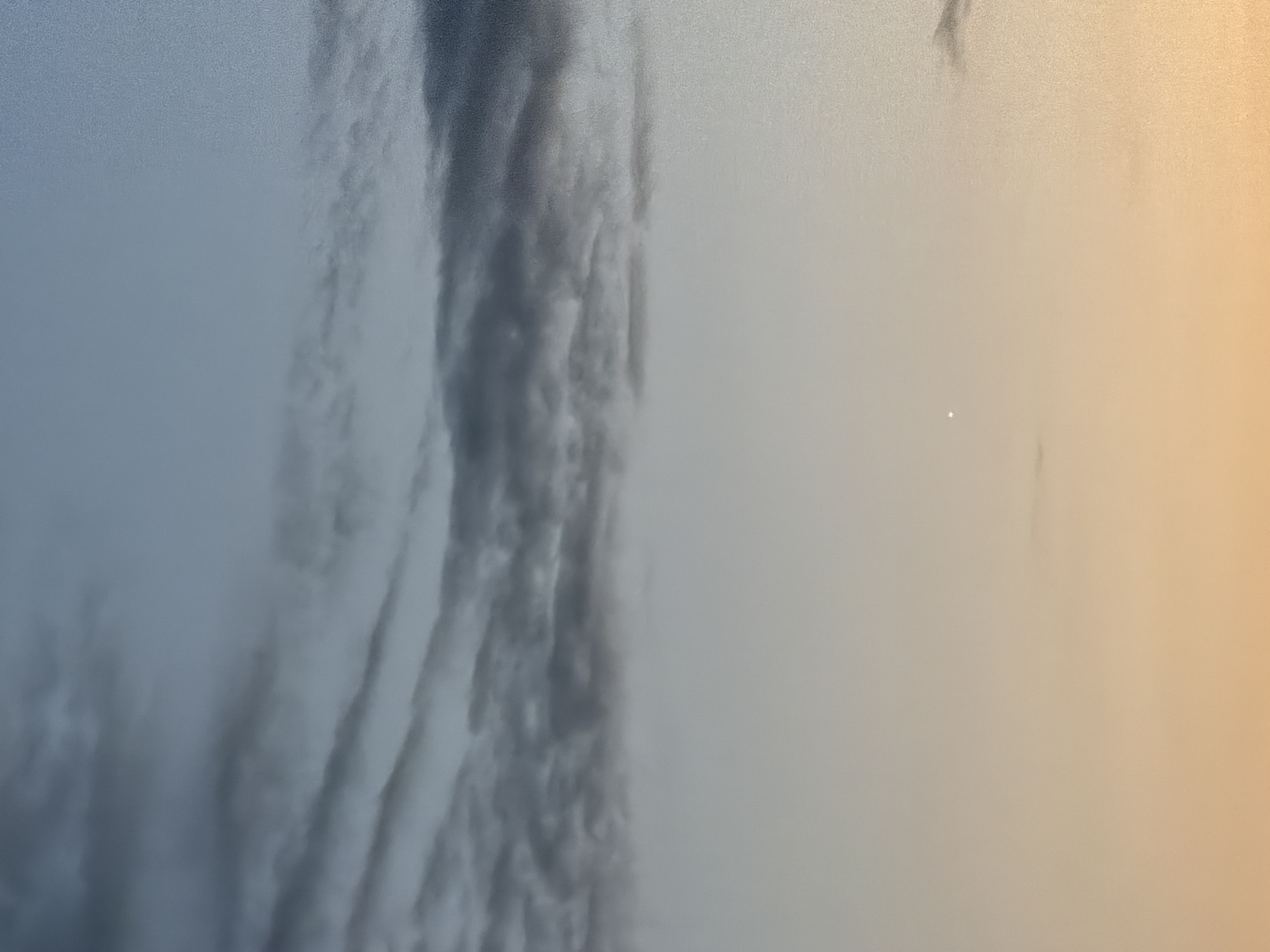 This is a close-up, eye-level, outdoor, medium-format photograph of a sky at dusk. The sky is layered with gradients of color, transitioning from a soft orange and yellow at the bottom to a pale blue at the top. Wispy, dark gray clouds are scattered across the upper portion of the image, appearing textured and somewhat elongated. A single, small, bright white speck, possibly a star or a planet, is visible in the lower-middle section of the frame, against the lighter colored sky. The overall impression is one of a serene, twilight sky. There are no people, animals, or specific landmarks visible to indicate a precise location within Kyiv, Ukraine.
