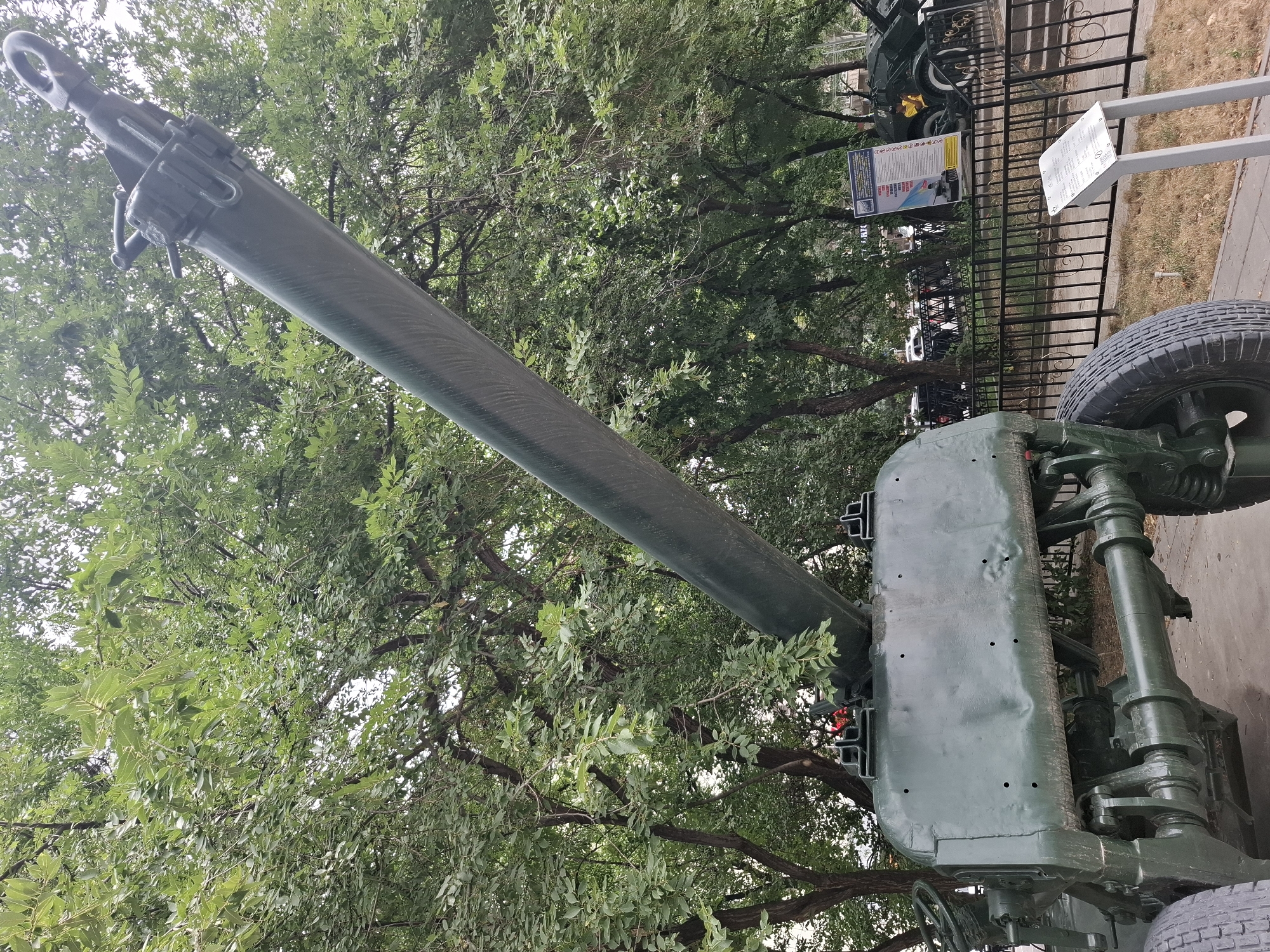 The image shows a close-up, low-angle view of a large, green, artillery piece, possibly a howitzer or cannon, positioned outdoors. The barrel of the weapon is prominently featured, extending diagonally upwards and to the left, set against a backdrop of lush green trees with dense foliage. The weathered green paint on the artillery piece shows signs of wear, including dents and small holes on the main body.
The artillery piece is mounted on a wheeled carriage, with one large, black tire visible on the right side of the frame. The complex mechanical components of the carriage, painted in the same dark green, are also visible.
In the background, behind a black metal fence, there are signs with text and images, suggesting this might be a museum exhibit or a memorial. One sign, in particular, has a blue and yellow color scheme with text in what appears to be Cyrillic script. Another sign is white with dark text. Beyond the fence, a paved walkway and some dry grass are visible, hinting at a park or outdoor display area. The overall lighting suggests it is daytime, and the abundance of leaves implies a season other than winter. The scene is static, with no visible people or immediate activity. The context provided places this scene in Almaty, Kazakhstan.