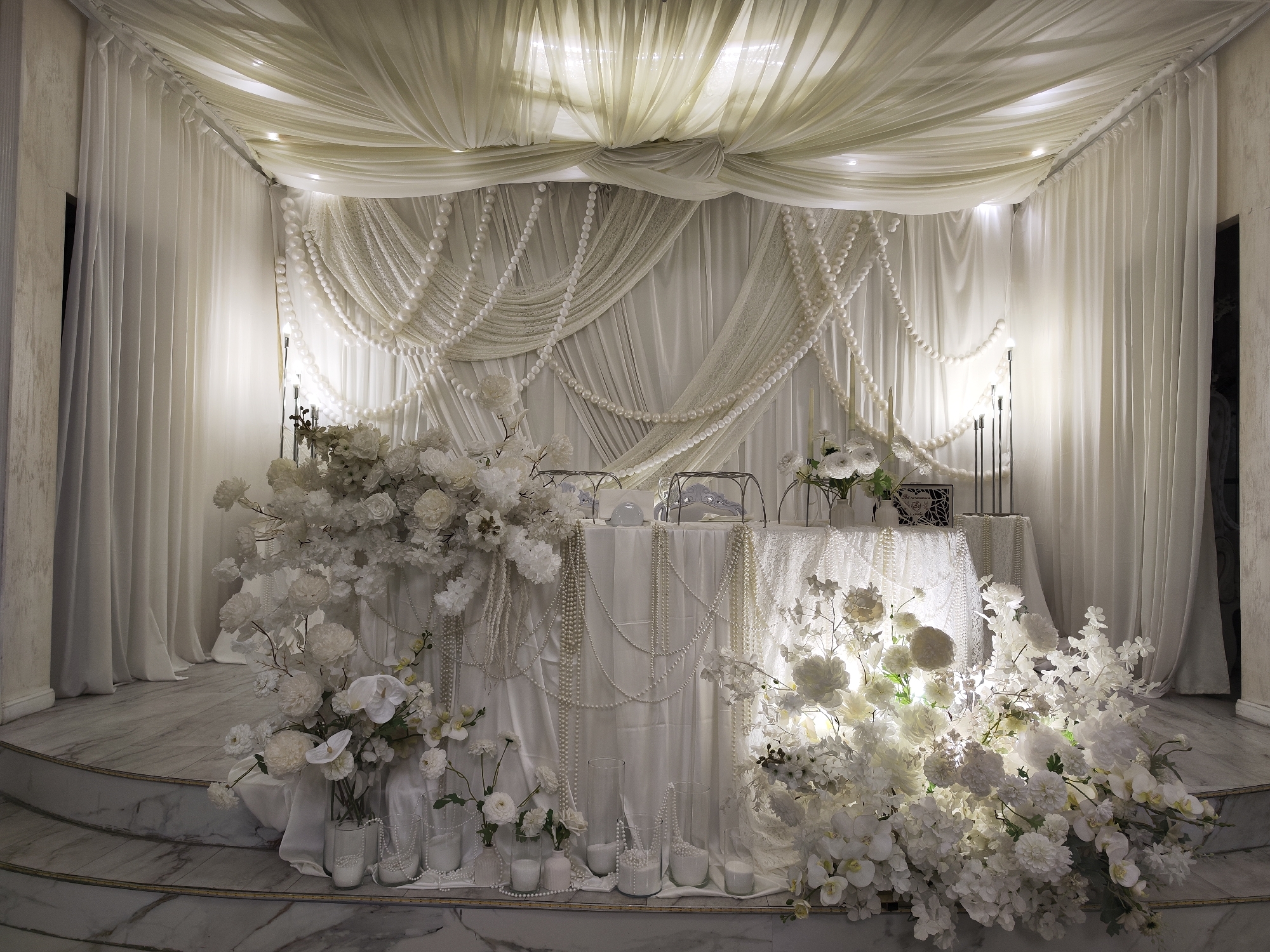The image depicts an elaborately decorated wedding setup in Vidnoye, Russia. The scene is dominated by white fabric drapery, cascading pearls, and abundant white floral arrangements. A draped ceiling with embedded lights creates a soft ambiance. The focal point is a long table, also draped in white fabric and adorned with more pearl strands. On the table, there are vases of white flowers, candle holders with dark candles, and decorative items. The table and surrounding area are set against a backdrop of sheer white curtains. The floor appears to be marble, with tiered steps leading up to the decorated platform. The overall aesthetic is luxurious and romantic, typical of a formal wedding reception or ceremony setting. There are no people visible in the image.