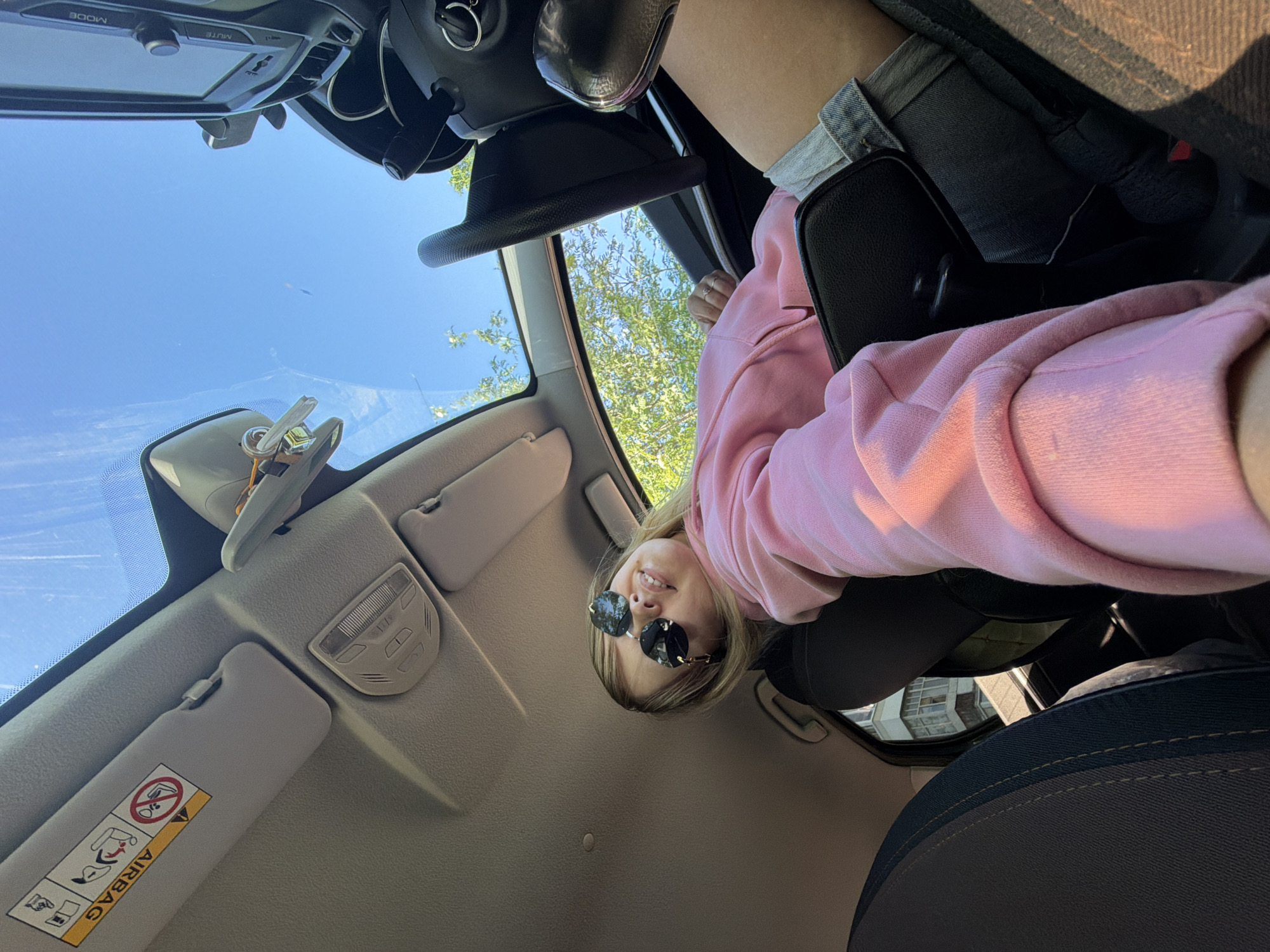 A fair-skinned woman wearing round sunglasses and a pink sweatshirt is inside a car. She looks down into the camera. The car's interior is beige, including sun visors, ceiling, and front dashboard. The steering wheel, seats, and center console are black. Outside the car's sunroof, a building is visible. The location in Chaikovsky, Russia is not determinable from this image.