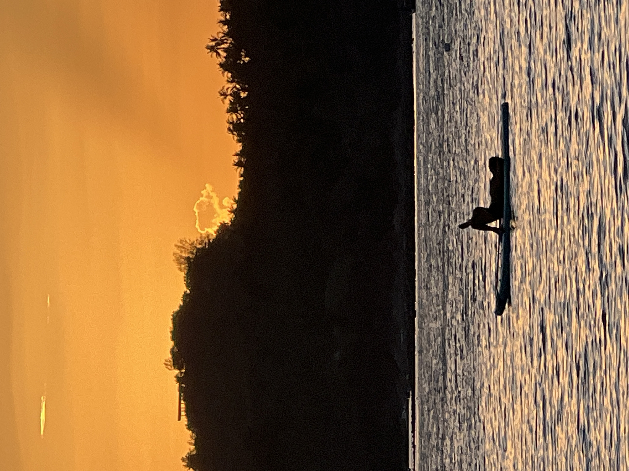 A silhouette of a person in a yoga pose is on a paddleboard in the ocean at sunset. The sun is setting behind a dark, forested hill, casting a warm orange glow across the sky and water. The person appears to be in a seated or reclined position, with their body curved. The water is calm, with gentle ripples reflecting the sunset colors. The hill in the background is covered in dense foliage and palm trees, with a small building visible on the ridge. The sky is a gradient of orange and yellow, with a few wispy clouds. The scene suggests a tranquil and serene moment at the end of the day. The location is the coast of Ko Pha-ngan District, Thailand.