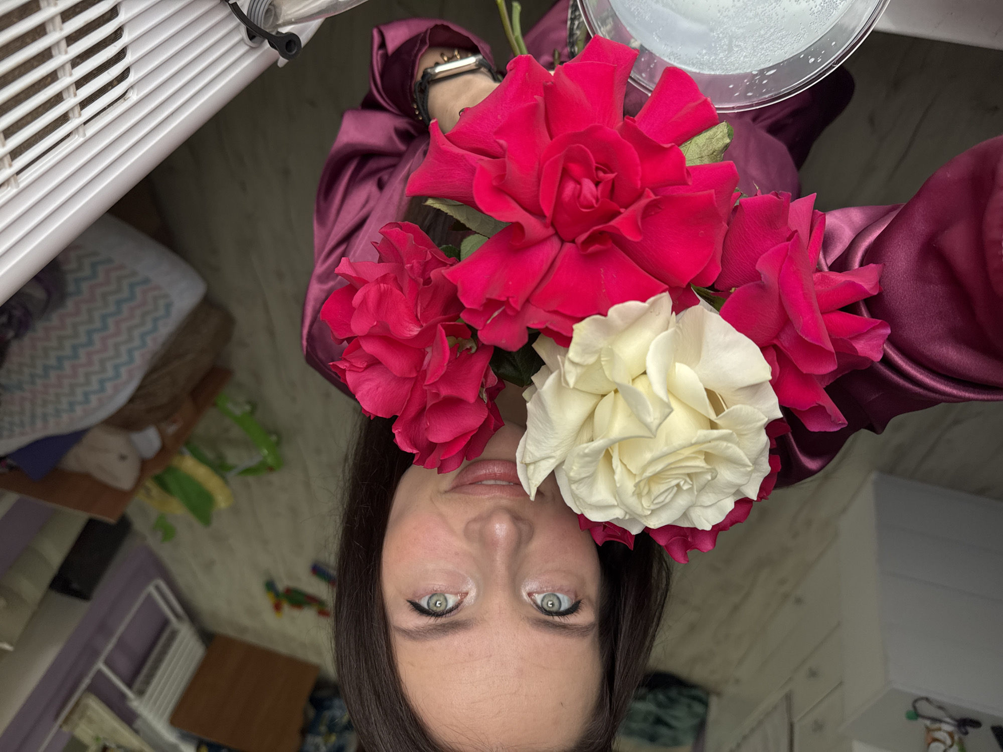 A woman with long dark hair and green eyes is holding a bouquet of red and white roses. She is wearing a satin burgundy dress. In the background, there are toys scattered on the floor of a room, suggesting a residential environment. The scene appears to be indoors. There is no indication of the specific location in Rusalevka, Russia.