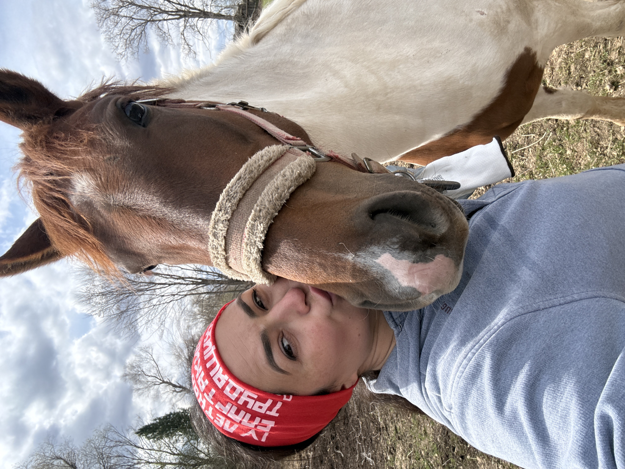 An intimate, low-angle shot captures a person and a horse engaging in a tender moment outdoors. The brown and white horse gently presses its muzzle against the person's cheek and shoulder, its halter visible. The person, positioned to the right, looks towards the camera with a subtle expression, wearing a light grey hooded sweatshirt and a red headband. The background features a partially cloudy sky and several bare trees, suggesting a late winter or early spring day in a rural or semi-rural setting, consistent with Кемуль, Russia. The red headband displays white Cyrillic text that reads "ТРУДАЙЛЯТ ТЕАТРЕТ," with the final "Т" in both words appearing inverted as part of the design. The interaction conveys warmth and affection between the human and animal.