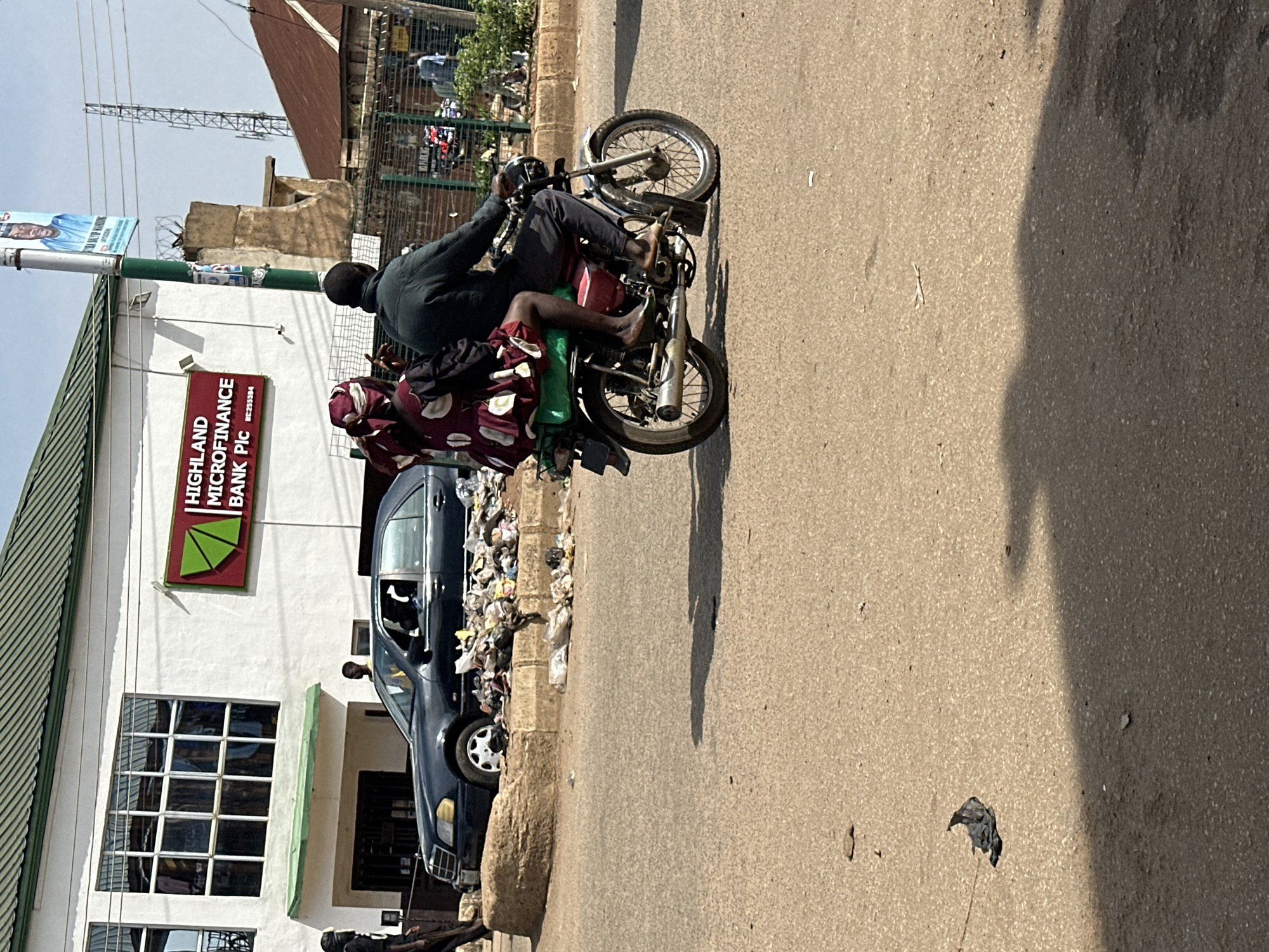 In Bukuru, Nigeria, a man and a woman ride a red motorcycle on a paved road. The woman, wearing a patterned dress and headscarf, sits behind the man, who is dressed in a dark jacket and trousers. The motorcycle has a green rack on the back, and its exhaust pipe is visible. In the background, a dark-colored car is parked near a white building with a green roof and a sign that reads "HIGHLAND MICROFINANCE BANK Plc." A pile of trash is next to the car. The environment appears to be a commercial area with a few people visible in the distance.