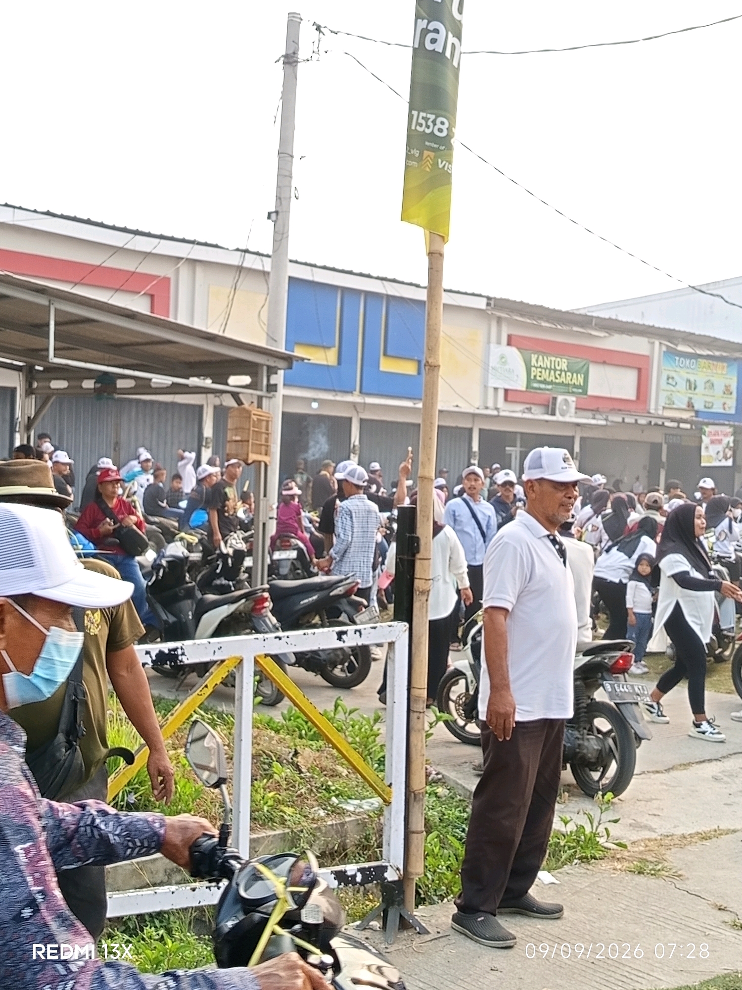 The image depicts an outdoor scene in the city of Karanganyar, Indonesia. In the foreground, a man wearing a white cap, white polo shirt, and brown pants stands facing right, his body in profile. To his left, another person wearing a white cap and blue face mask is partially visible. Behind them, a crowd of people, many wearing white caps or head coverings, are gathered, some near motorcycles. Several motorcycles are parked in the scene, and a building with signage is visible in the background. The environment appears to be a street or public area, with a sidewalk, a pole with wires, and vegetation. A banner on a pole in the center of the image has Indonesian text. The timestamp on the bottom right of the image indicates "09/09/2026 07:28".