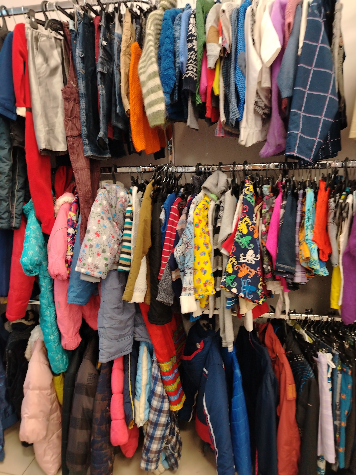 The image shows a rack of children's clothing in a store in Yaroslavl, Russia. The clothing includes jackets, pants, and sweaters in various colors and patterns. The clothes are hung on hangers on a metal rack. The background is blurred, but it appears to be a store interior with other racks of clothing.