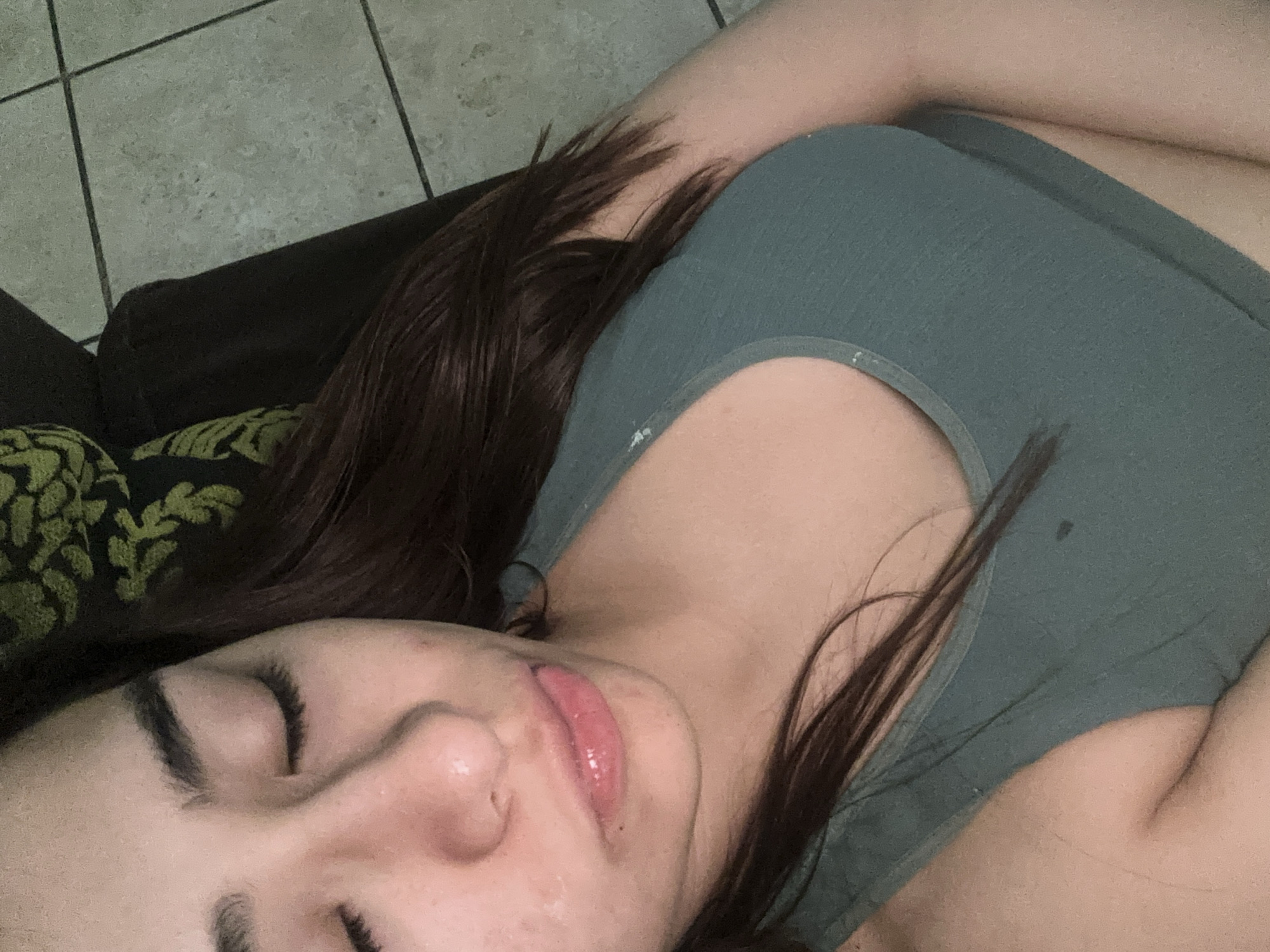 A woman is reclined on a cushioned surface, possibly a couch or a bed, with her eyes closed and hair spread out. She wears a green top. The background shows a tiled floor and part of the furniture. The location cannot be determined from the image alone, and there is no indication that it is Puebla, Mexico.