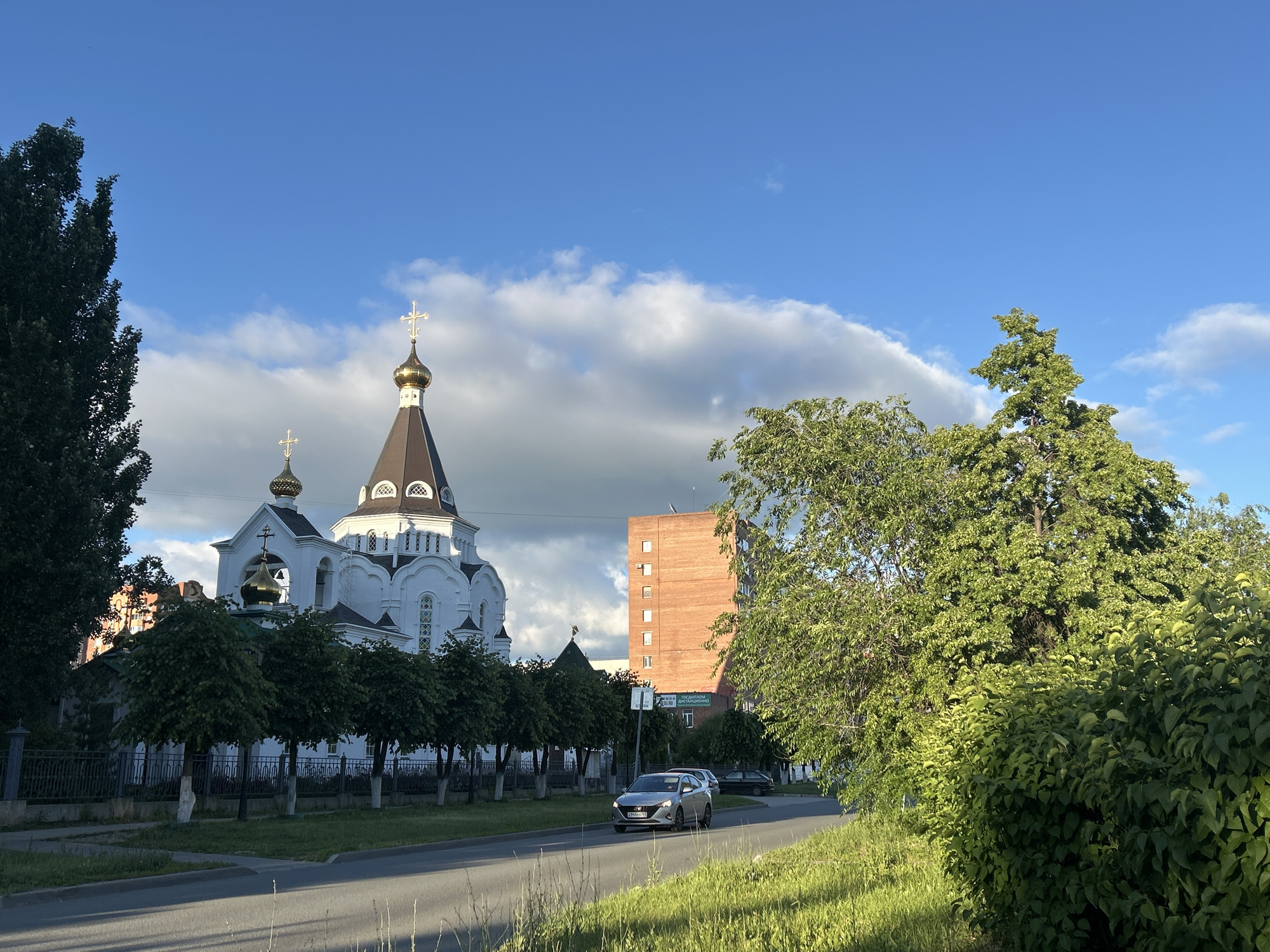This image presents an urban landscape in Tolyatti, Russia, featuring a prominent white Orthodox church under a bright blue sky with scattered white clouds. The church, situated on the left side of the frame, boasts a dark grey roof and several golden onion-shaped domes, each topped with a golden Orthodox cross. A smaller bell tower with a similar dome and cross is also visible.
In front of the church, a row of neatly trimmed green trees stands behind a dark metal fence, separating the religious complex from the road. To the right of the church and further down the street, a tall, multi-story red brick building with uniformly placed windows is visible. A sign affixed to a pole near this building displays Russian text, partially decipherable as "ПОДГОТОВКА ДИСТАНЦИОННО" (meaning "Distance Learning/Preparation").
An asphalt road runs horizontally across the lower part of the image. A silver sedan is seen driving on this road, heading away from the viewer. The car's license plate shows "895АХ 163," which is a Russian plate, with '163' indicating the Samara Oblast, where Tolyatti is located. The foreground on the right features abundant green foliage, including tall trees and dense bushes, which cast long shadows, suggesting that the photograph was taken during the late afternoon or early evening. The overall scene blends traditional religious architecture with everyday urban life and natural elements.
