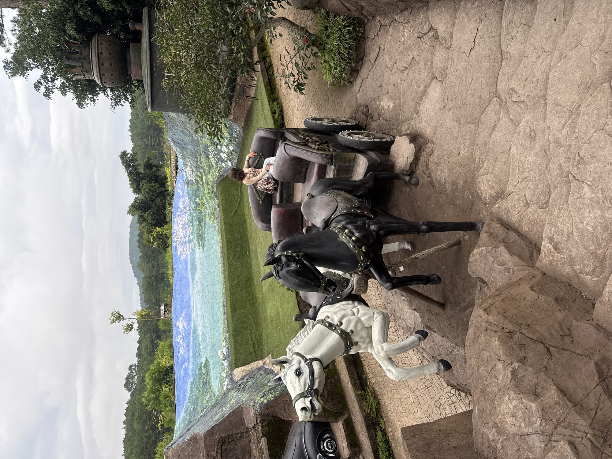 This is a full shot of a decorative carriage pulled by two horse statues in front of a painted mural depicting a natural landscape. The carriage is dark brown with a seated figure wearing a light-colored outfit and a patterned scarf. The horses are one white and one black, with detailed harnesses and saddles. Behind the carriage and horses, a large mural shows a body of water, green hills, and trees, with a cloudy sky above. The foreground features large, textured rock formations and a paved pathway. The scene appears to be part of an outdoor attraction or park in Da Lat, Vietnam, potentially a tourist spot designed for photo opportunities.