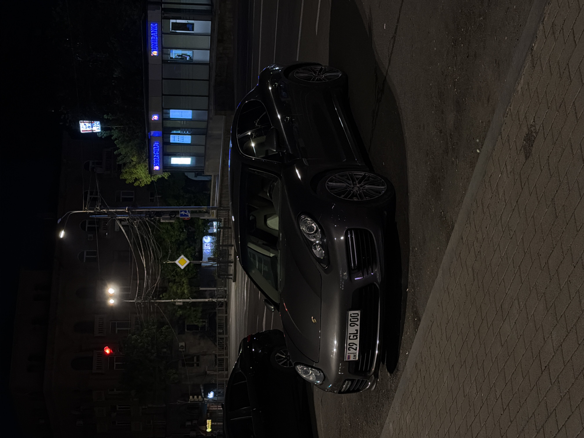 The image depicts a dark grey Porsche SUV parked on a street at night in Yerevan, Armenia. The car's front is facing the viewer, with its license plate "29 GL 900" clearly visible. A second, darker car is partially visible behind the Porsche. In the background, there's a Unibank building illuminated with blue signage. Streetlights are on, and a traffic light shows red. The surroundings appear to be urban, with buildings, trees, and street furniture visible in the distance. The street is paved, and a brick sidewalk is to the right of the vehicle.