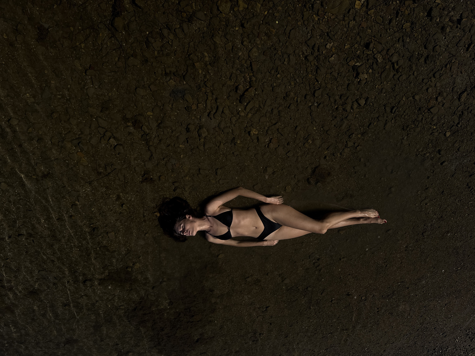 A woman in a black bikini is lying on her back in shallow, dark water. Her eyes are closed and her head is tilted back. Her hair is spread out around her head. Her legs are crossed at the ankles. The bottom of the water is rocky and dark. There is no indication of the location in Mrčevac, Montenegro.