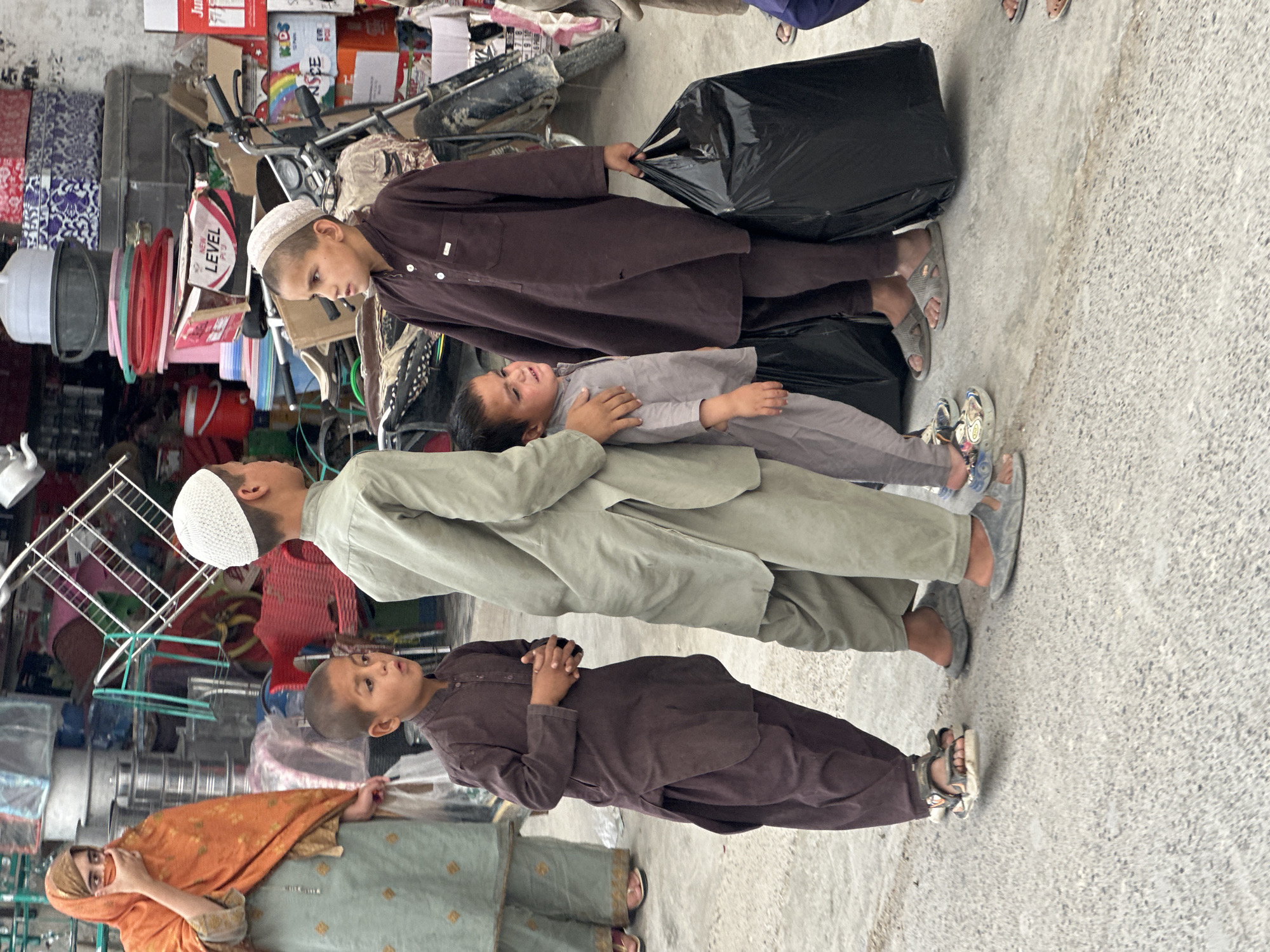 The image shows four children and a woman standing on a concrete surface. The location appears to be a street, evidenced by the presence of merchandise and possibly storefronts in the background.
The woman is wearing an orange headscarf and a blue dress. Three of the children are wearing brown, green, and grey traditional clothing, respectively, while the other child is holding a black trash bag. The three boys wear white caps. The smallest child wears a grey outfit and lays across the boy in green. The objects in the background appear to be general merchandise, including kitchenware, tools, and other household items. One child is holding a large, black garbage bag.