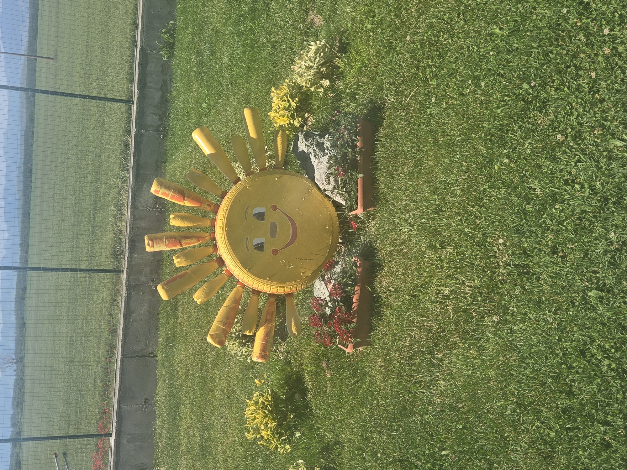 A grassy lawn features a sun decoration. The sun is yellow with orange accents on its rays. The lawn is vibrant green and bordered by a concrete strip next to a chain-link fence. Behind the fence, there's a view of distant trees and a mountain range under a blue sky. Adjacent to the sun, there are plants in terracotta pots. Location: Unknown, Unknown.