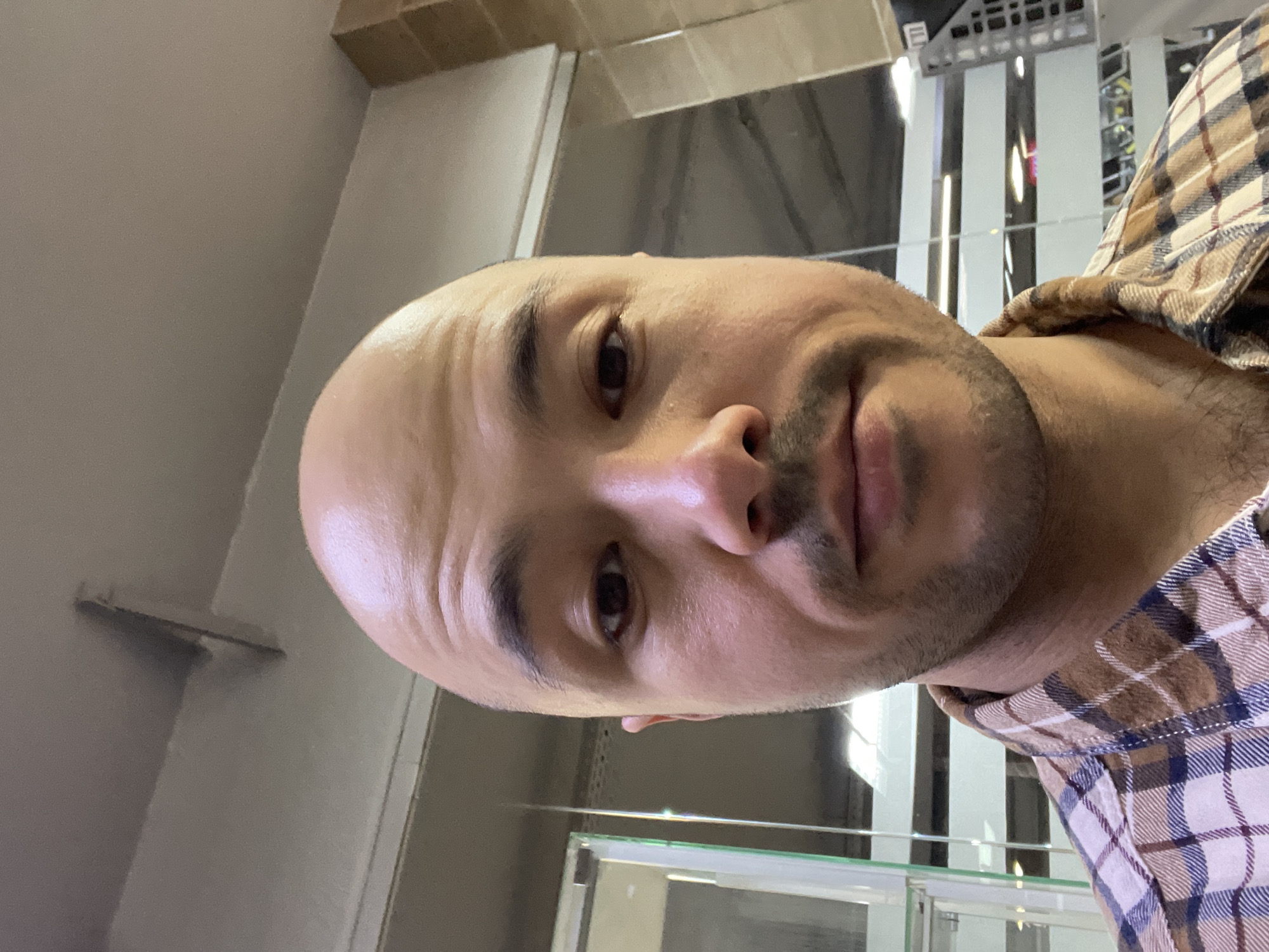 A close-up, slightly angled headshot depicts a bald, light-skinned man with a trimmed beard. He has dark eyes and is wearing a tan and blue plaid shirt. The background includes blurred features suggesting a modern interior with reflective surfaces, possibly a commercial space or retail environment. The location is unknown, I cannot identify the location as Izhevsk, Russia.
