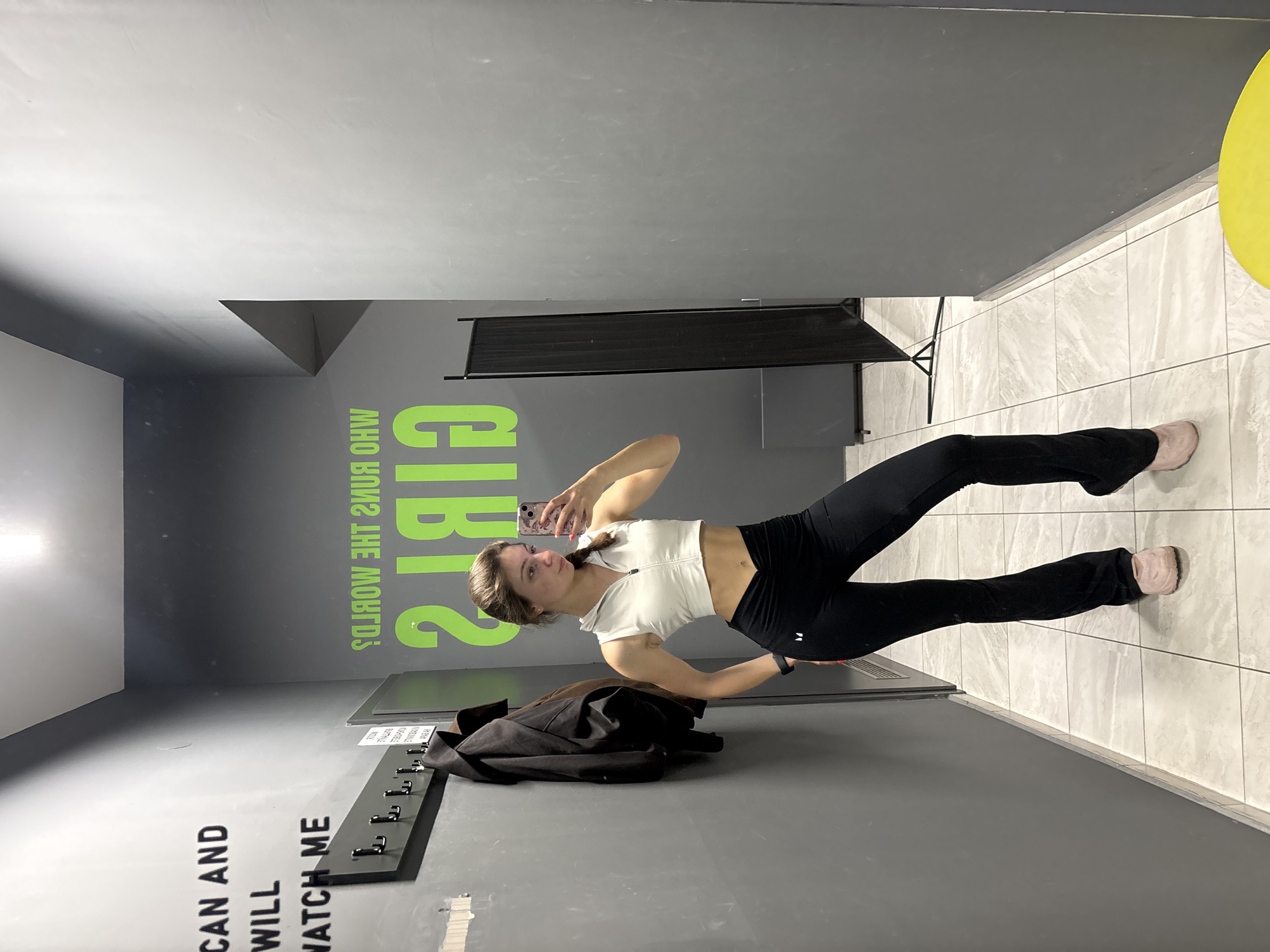 A young woman is taking a selfie in what appears to be a changing room. She is wearing a white athletic crop top and black leggings. Her hair is in a ponytail. A black jacket is draped over a coat rack on the wall to her left. Text reading "WHO RUNS THE WORLD GIRLS" is written on the wall behind her. The room has gray walls and a tiled floor. A black privacy screen is positioned in the background. It is impossible to determine if the location is in Varna, Bulgaria based solely on the provided image.