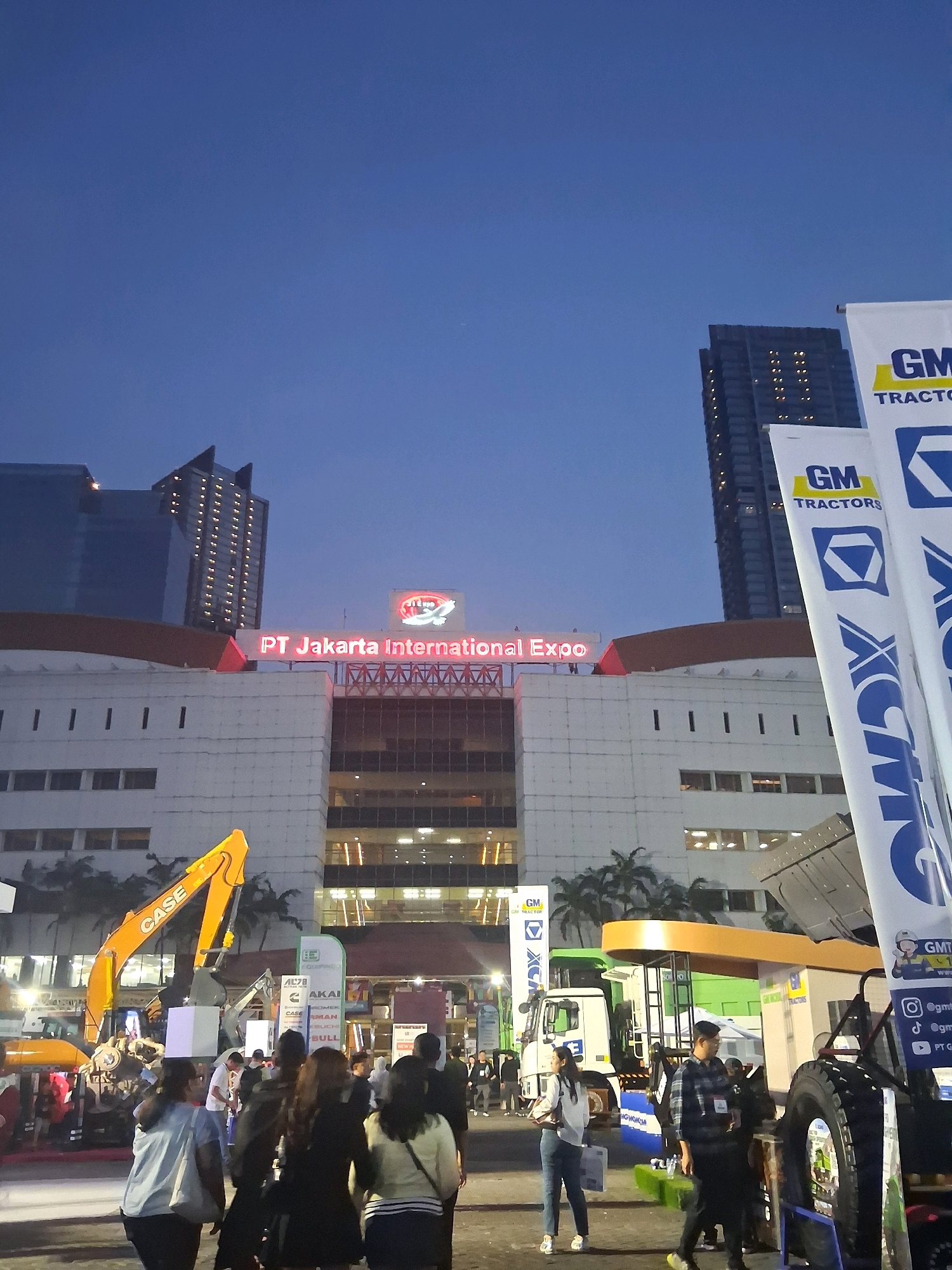 The image captures a scene at the PT Jakarta International Expo, located in Central Jakarta, Indonesia, during what appears to be the evening or twilight hours. The sky is a deep, clear blue, transitioning into darker shades as it nears the horizon. Prominently featured is the facade of the exhibition center, a large, multi-story building with white and light-colored walls. Above the main entrance, a sign in bright red neon letters reads "PT Jakarta International Expo". Several tall, modern buildings, including skyscrapers, are visible in the background, some with illuminated windows, suggesting the surrounding urban environment.
In the foreground, there is a bustling atmosphere with a group of people milling about. Attendees are dressed in casual clothing. Some individuals are walking, others appear to be conversing. Among the subjects, there are several large pieces of heavy machinery, specifically construction vehicles, displayed as part of the exhibition. A bright yellow excavator with the brand "CASE" is particularly noticeable on the left side of the image. Other vehicles, including trucks, are also present. Tall white banners with blue and yellow branding, including the logo "GM TRACTORS", are erected, indicating exhibitors. The lighting appears to be a mix of ambient twilight and artificial lighting from the exhibition displays and surrounding buildings. The overall impression is that of a lively event taking place within a significant urban exhibition venue.