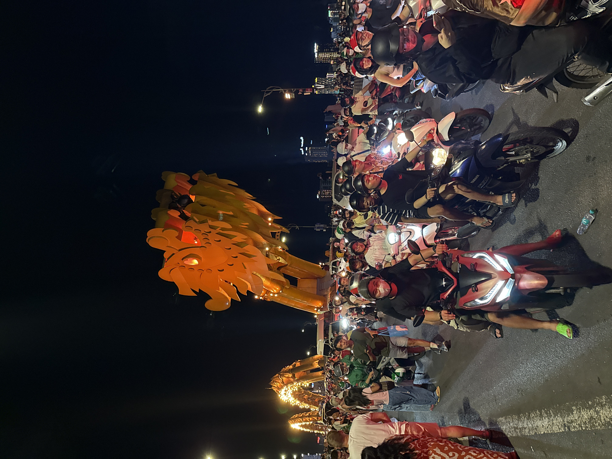 This image captures a dense nocturnal scene on the Dragon Bridge in Da Nang, Vietnam. The prominent, illuminated golden head of the Dragon Bridge occupies the upper right portion of the frame, with its intricate scales and glowing red eyes clearly visible. Further along the bridge to the left, several illuminated golden arches, representing the body of the dragon, are also visible.
The road is heavily populated by a multitude of individuals, predominantly of Asian descent, riding or standing beside motorbikes. The density of people and vehicles suggests either a significant traffic jam or a crowded gathering. Most people on motorbikes are wearing various types of helmets, and some individuals are also wearing face masks.
In the immediate foreground, a person on a red Honda motorbike with illuminated headlights is prominent, wearing a black shirt, a red helmet, and green flip-flops. Another individual on a blue Yamaha motorbike is visible further left. Several other motorbikes, including white, black, and various colored models, are packed tightly across the visible road surface. Headlights and taillights of vehicles contribute significantly to the scene's illumination.
On the left side of the frame, several people are standing or walking on the roadside, including an individual in a white t-shirt and another in a red patterned outfit. A person in a green jacket is visible amongst the motorbikes on the left. In the mid-ground, a person on a motorbike is actively using a mobile phone.
The asphalt road surface shows some reflective patches and faint white markings. Overhead streetlights emit light onto the scene. In the background, beyond the immediate crowd, urban buildings with illuminated windows are visible against the dark night sky, indicating a city environment. A discarded plastic bottle lies on the road surface in the lower left. The overall scene is brightly lit by artificial sources against the dark backdrop of the night.