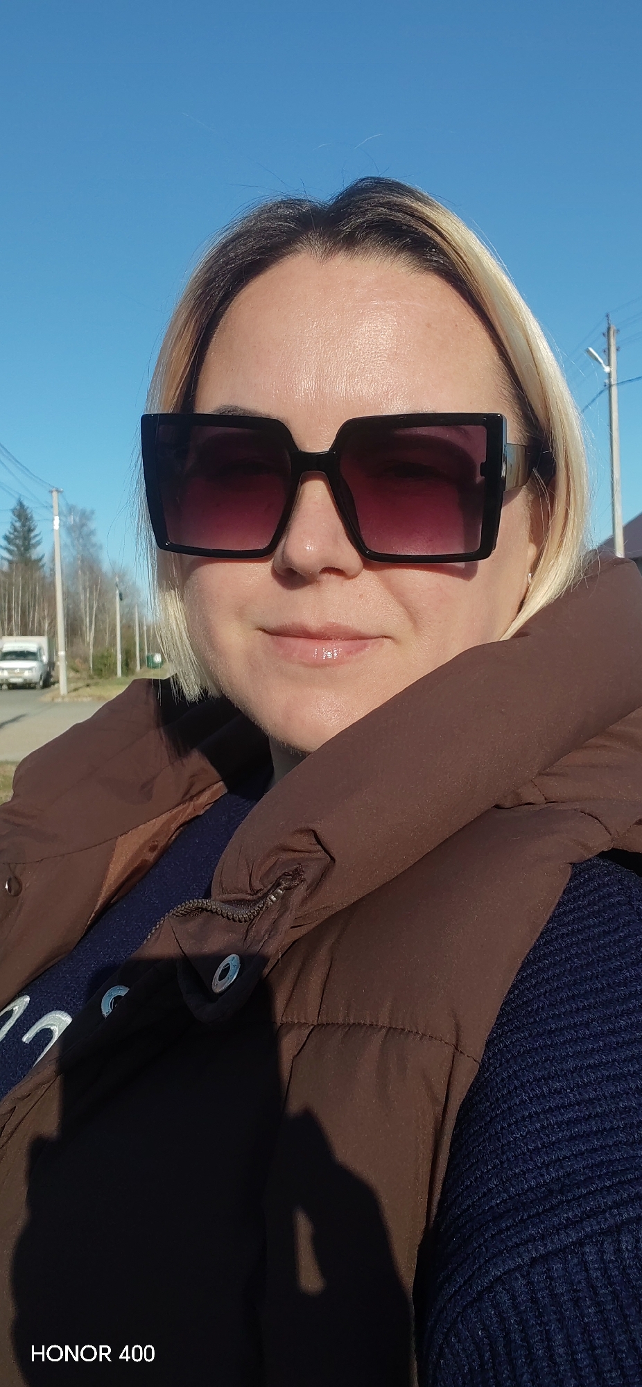 A woman is taking a selfie outdoors. She is wearing oversized square sunglasses, a brown puffer vest, and a blue sweater. Her hair is blonde. In the background is a road, trees, a white van, and power lines. The image is marked "HONOR 400". There is no visible information that indicates the location is the city of Noviy, Russia.