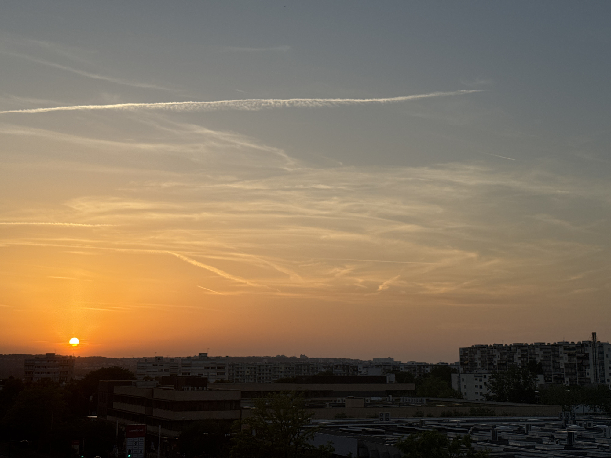 A wide shot captures an urban skyline at sunset. The sun, a bright orange orb, is partially obscured by a building to its left and is low in the sky, casting a warm glow across the scene. The sky transitions from a vibrant orange at the horizon to a pale blue at the top. Wispy clouds, some appearing as contrails, are scattered across the sky. In the foreground, a dense collection of buildings creates a layered urban landscape. Some buildings are taller and modern, featuring numerous windows and air conditioning units, while others are lower and appear to be industrial or commercial structures. Trees are visible, adding touches of dark green to the scene. The overall impression is one of an ordinary city at the end of the day, bathed in the colors of sunset.
