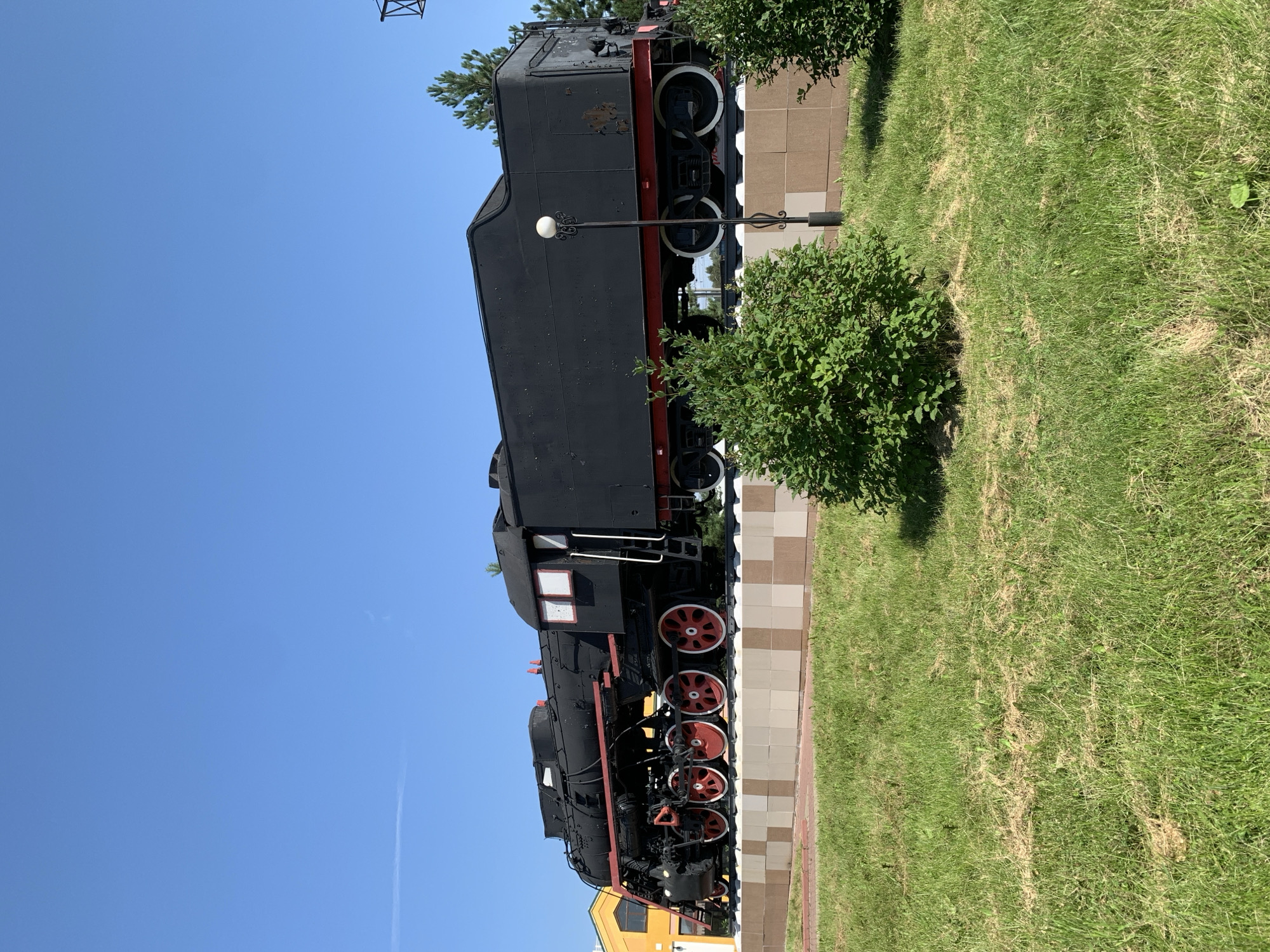 A black steam locomotive, likely a monument, is displayed outdoors on a platform. The locomotive is positioned on tracks and appears to be a static exhibit. The platform is made of tiled blocks in shades of beige and brown. In front of the platform, there is a well-maintained green lawn with some dry patches and scattered leaves. Two lush green bushes are situated in the foreground, partially obscuring the base of the platform. A lamppost with a decorative black metal base and a white globe-shaped light is visible behind the bushes and in front of the locomotive. The background features a bright blue sky, a section of a yellow building with windows on the left, and some distant trees and foliage. The scene is illuminated by daylight, suggesting a clear and sunny day.
The location is identified as Prokopyevsk, Russia, a city known for its industrial history, particularly coal mining. The presence of a steam locomotive monument is fitting for a city with a significant railway presence related to its industrial activities.