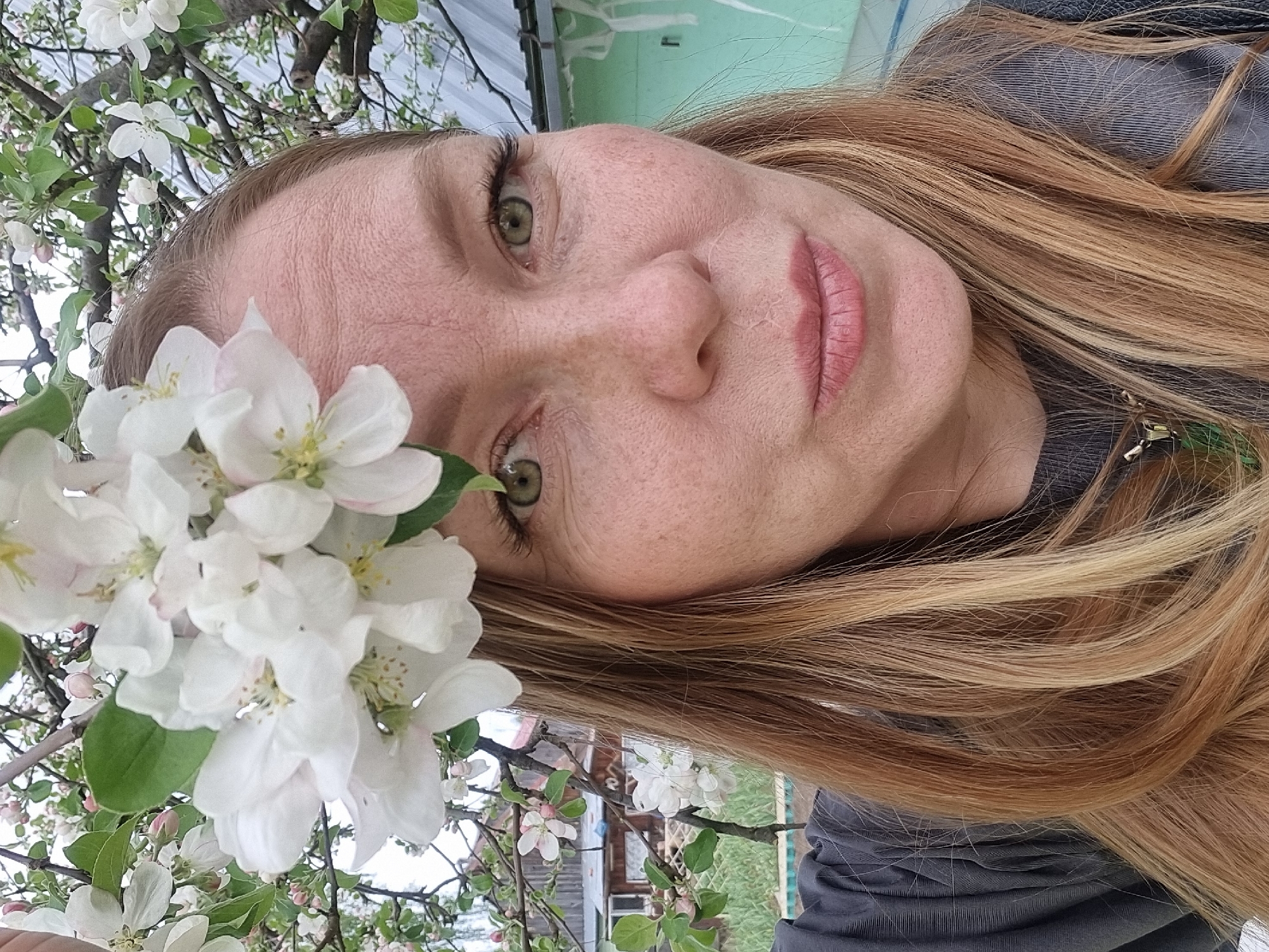 A fair-skinned woman with freckles and light brown hair, partially obscured by white blossoms, is photographed in an outdoor setting. Her face is angled towards the camera, revealing green eyes and light pink lips. She is wearing a grey shirt. The background reveals flowering branches of what appears to be a blossoming tree. Distant elements suggest a residential area, potentially in the city of Chaikovsky, Russia, characterized by foliage, a building structure, and fencing. The scene is captured in daylight.