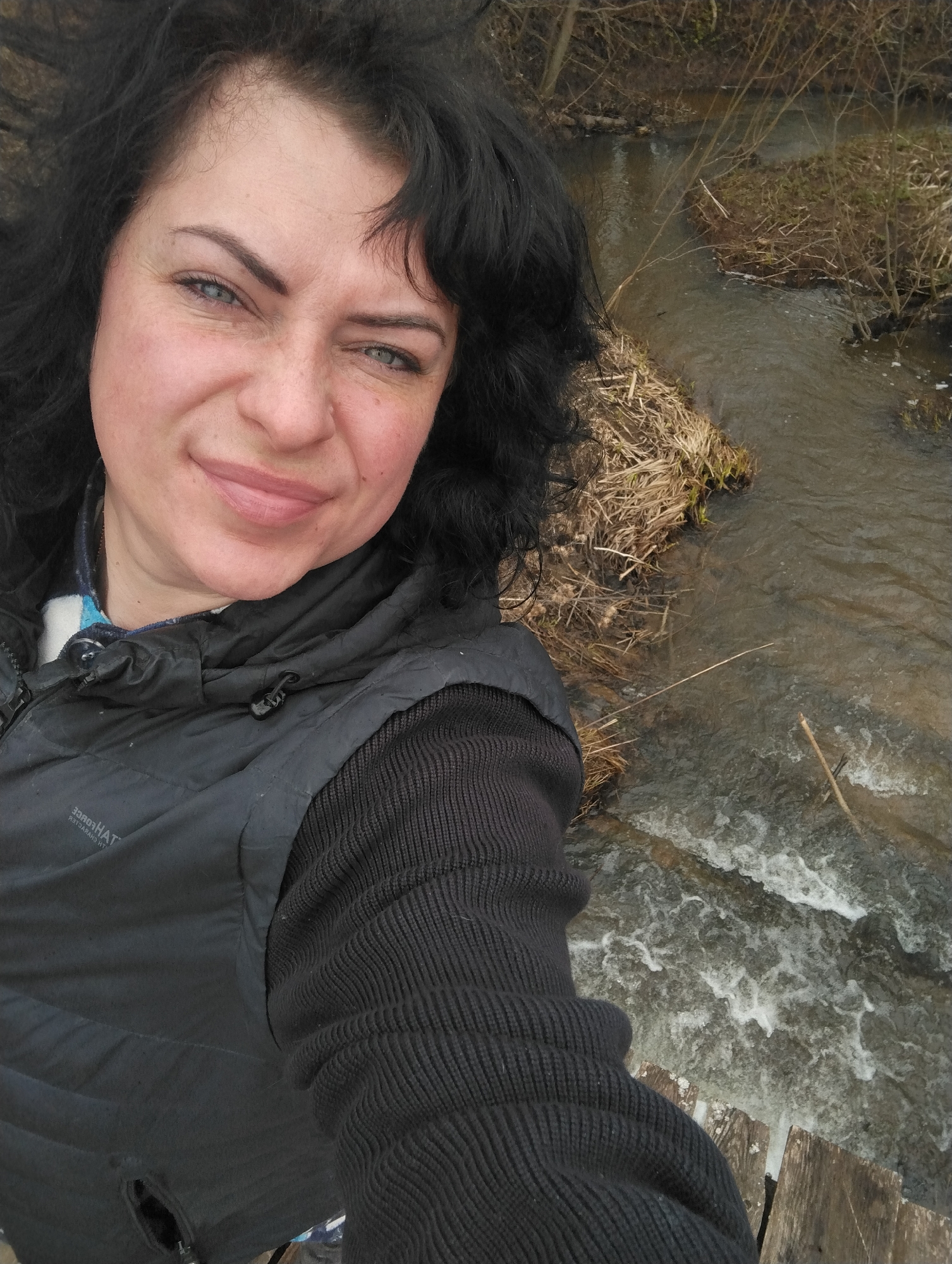 A woman with dark, curly hair, light skin, and blue-green eyes looks directly at the camera, taking a selfie outdoors in Grishanki, Russia. She has a subtle smile and crinkled eyes, conveying a pleasant expression. She is wearing a dark, possibly black or dark grey, quilted vest over a ribbed, dark long-sleeved top, with a blue and white patterned collar visible underneath. On the vest, inverted text reads "3DЯOТHАТ ЯЭТГАЯАЯЧ ИН" (likely "HATHRORD CHARGERS" mirrored).
The setting is a natural, outdoor environment, with the woman positioned above a flowing stream or small river. The water below her is choppy with whitecaps in the immediate foreground, transitioning to calmer sections further back. The banks of the stream are covered with dry, brown grass and sparse, bare tree branches and bushes, indicating a late winter or early spring season. A small wooden structure, possibly part of a bridge or walkway, is visible at the bottom right. The overall perspective is a high-angle shot looking downwards, capturing both the woman and the natural landscape.