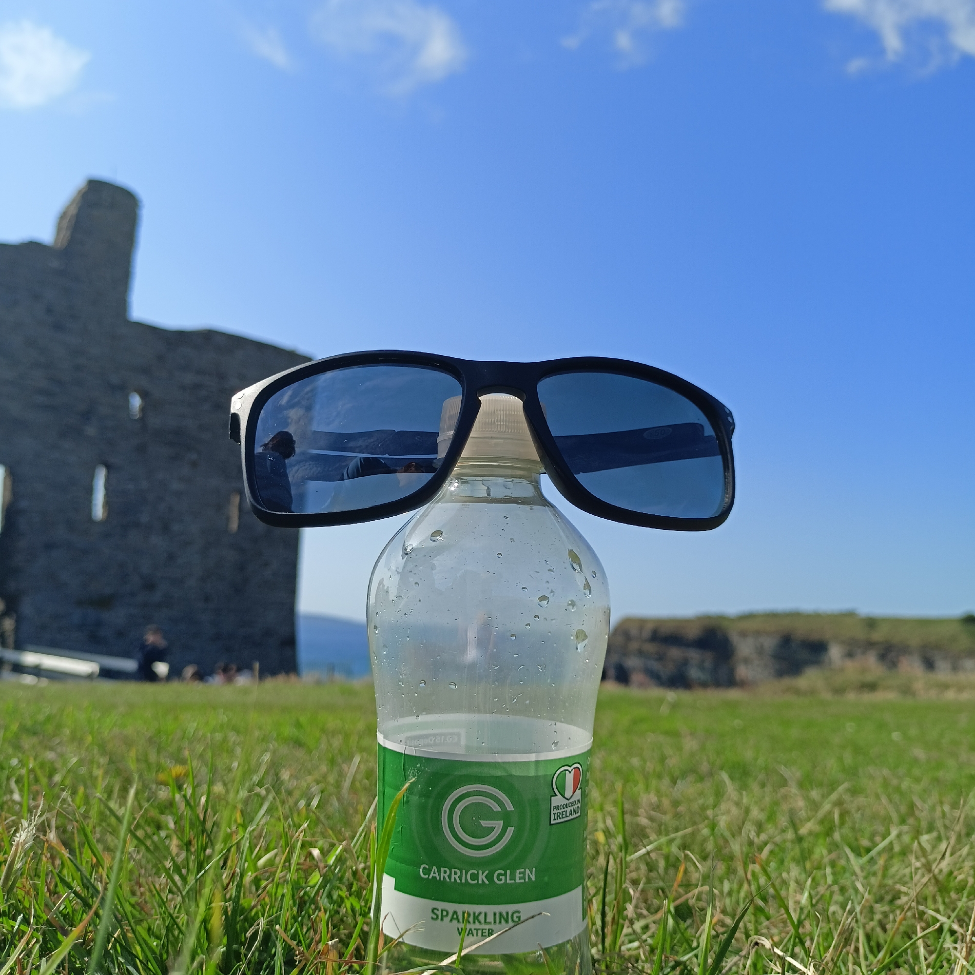 An outdoor, low-angle, medium shot captures a clear plastic bottle of Carrick Glen sparkling water placed on a grassy knoll, with a pair of black sunglasses perched on top of its cap, facing the camera. The bottle's label, featuring a green circle with a white 'G' and the text "CARRICK GLEN SPARKLING WATER," is clearly visible. A small "PRODUCED IN IRELAND" emblem with the Irish flag is also present. The background is a bright blue sky with scattered white clouds. To the left, a blurred, ancient stone structure, resembling a castle ruin, dominates the middle ground. In the far background, a rocky coastline is visible, suggesting a coastal location. The overall scene is brightly lit, indicating daylight. The approximate location is Ballybunnion, Ireland.
