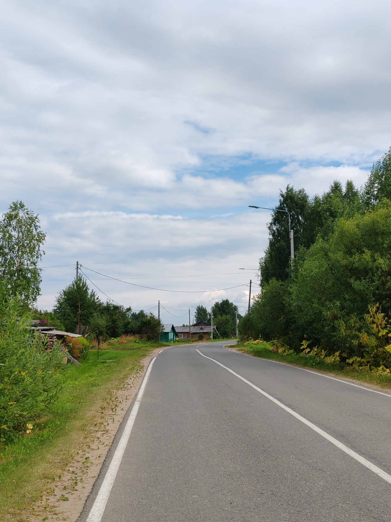 The image shows a paved road with a white line down the center, curving to the right. The road passes through a rural area with trees and vegetation on both sides. On the left side of the road, there are some buildings and a pile of wood. On the right side of the road, there are more trees and some yellowing foliage. In the distance, there are a few houses with green roofs. The sky is cloudy with patches of blue. The scene appears to be a typical village road in Russia.