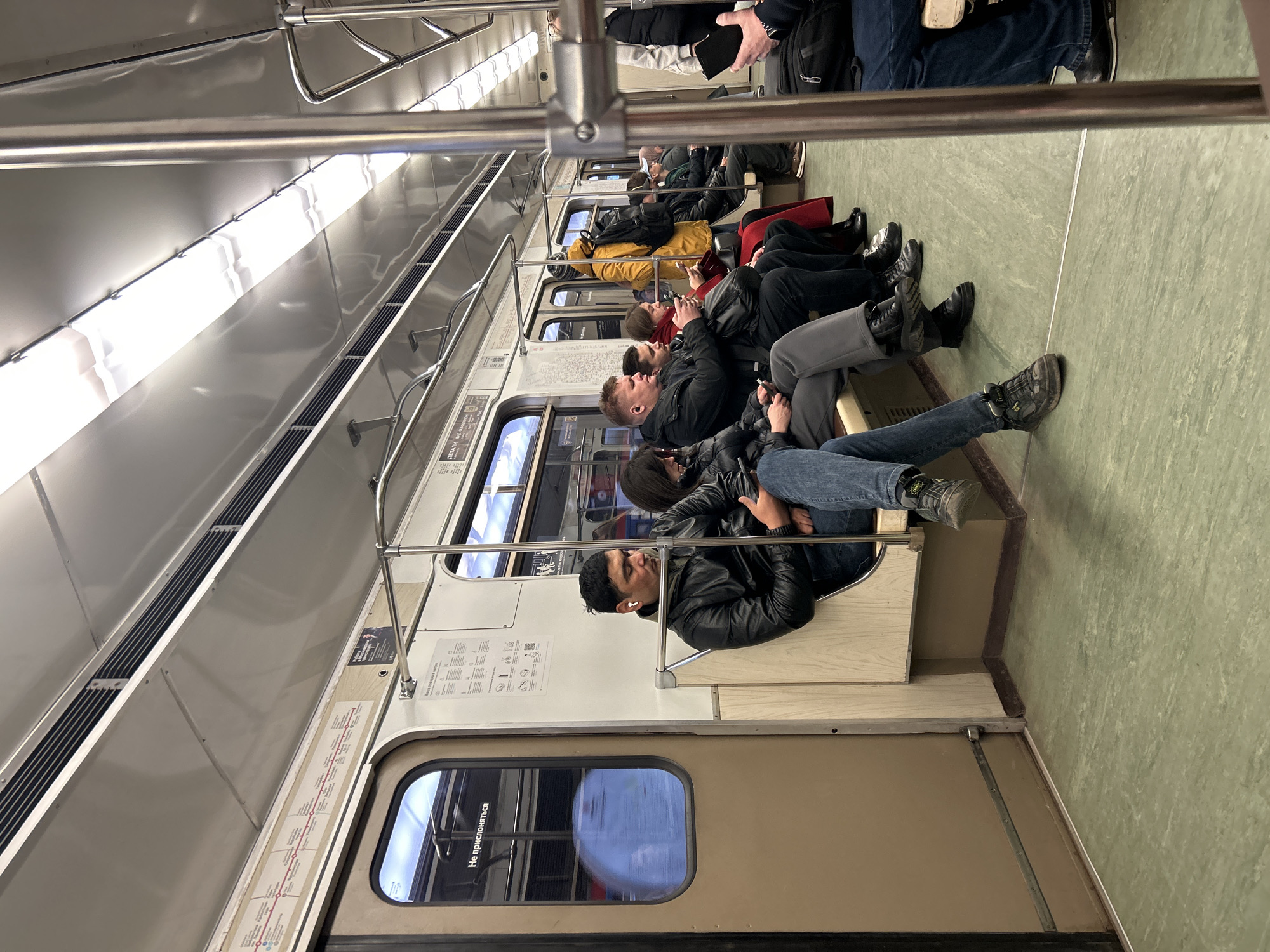 The image shows the interior of a metro train car, populated by numerous passengers. The perspective is from within the carriage, looking down the aisle at rows of seated individuals. The image is oriented vertically, suggesting it was taken with the camera rotated 90 degrees.
**Subjects:** The primary subjects are the passengers, mostly adults, who are seated on beige-colored bench seats. Many are dressed in winter attire, including jackets and coats in various colors like black, yellow, and red. Several individuals are engrossed with their smartphones. One man in the foreground, wearing a black leather jacket and white earbuds, appears to be resting or sleeping. Other visible objects include metal handrails and support bars running along the ceiling and between seat sections, overhead fluorescent lighting fixtures, and dark ventilation grilles. The floor is covered with a speckled green material.
**Activity/Event:** The scene depicts people commuting or traveling by metro. They are engaged in typical passenger activities such as sitting, observing, using mobile devices, or resting.
**Notable Details:** The setting is definitively inside a public transport train, specifically a metro, consistent with the provided context of Moscow, Russia. The interior design, including the type of lighting, handrails, and seating, is characteristic of older metro systems. No specific time of day is discernible, as the primary light source is artificial. Emotions appear neutral, reflecting the common experience of public transport.
**Visible Text:** Russian text is visible on the windows and information panels. On the window, the phrase "Не прислоняться" (Ne prislovyat'sya), meaning "Do not lean," is clearly legible. A map strip along the upper wall section displays a metro line diagram with station names written in Cyrillic, further confirming the location. Other panels contain additional Cyrillic text, likely operational information or advertisements.