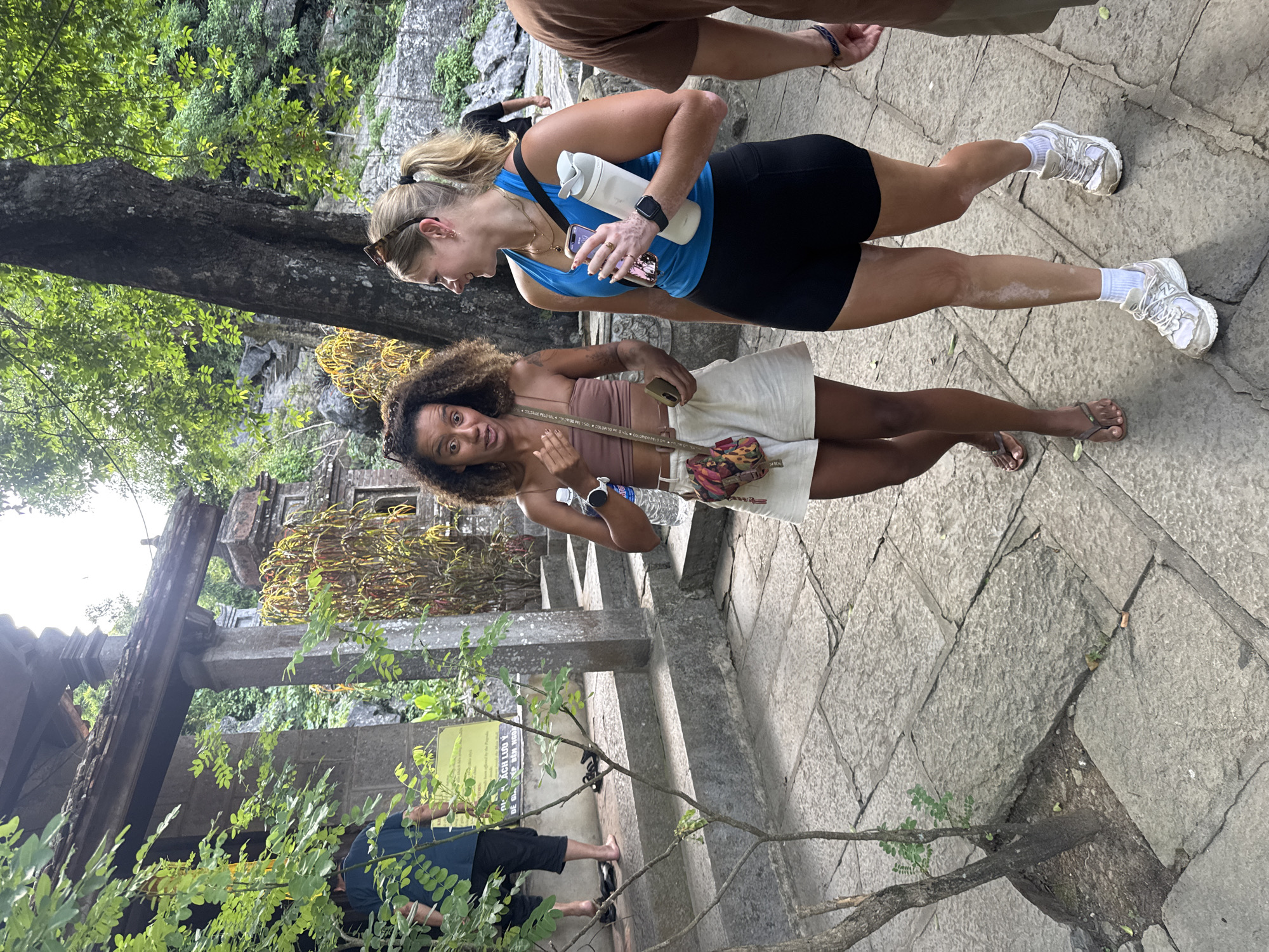 Two women are walking on a stone-paved path near a temple structure in Nam Hoa Lư Ward, Vietnam. The woman on the left has curly brown hair and is wearing a brown crop top and white shorts. She is holding a water bottle and a phone. The woman on the right has blonde hair pulled back in a ponytail and is wearing a blue tank top and black shorts. She is holding a white water bottle and a phone. They are walking towards the right of the frame. Behind them, there are stone steps, trees, and part of a temple building with a tiled roof. The ground is made of large, weathered stone slabs.