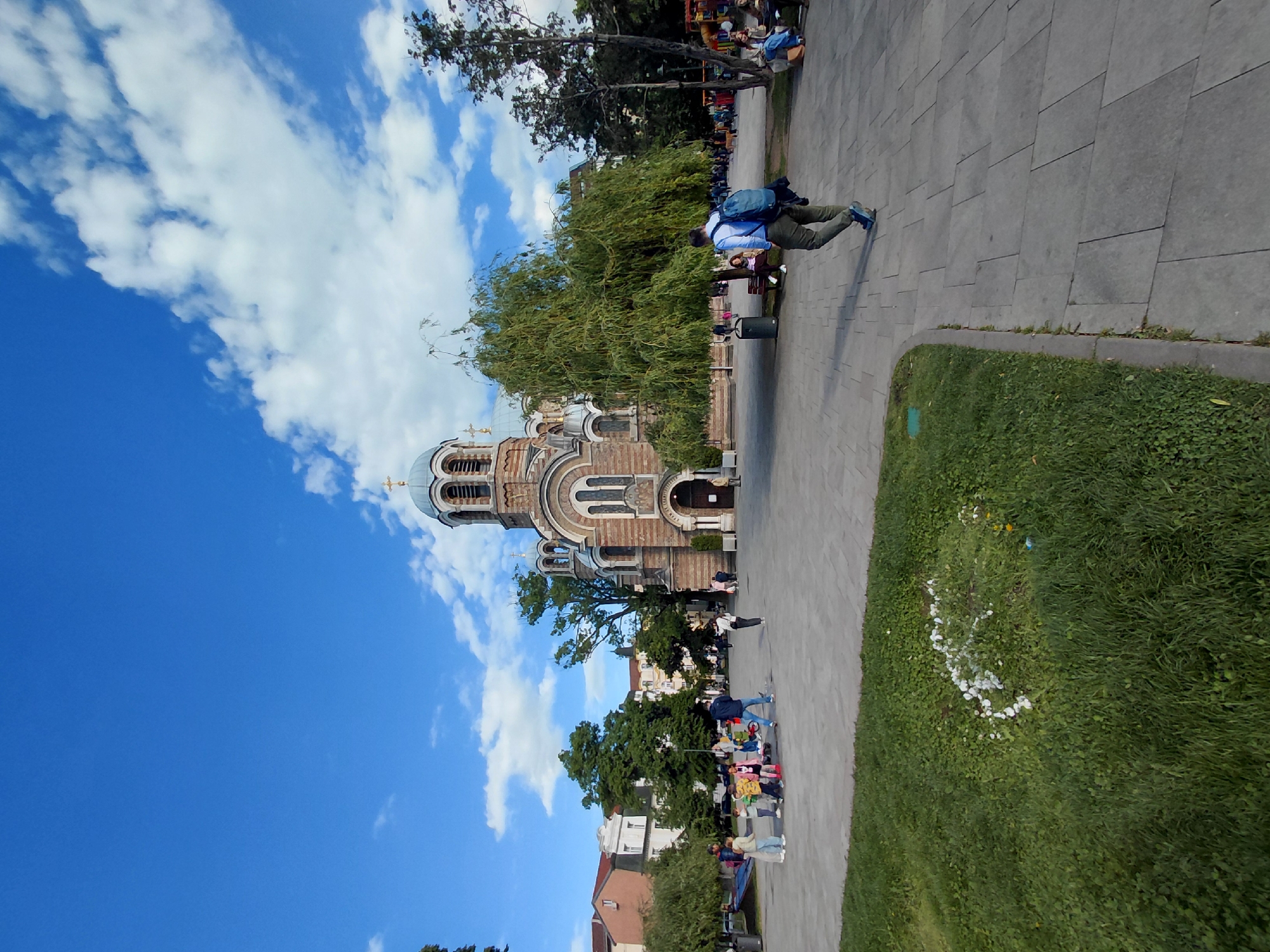 The image captures the Saint Alexander Nevsky Cathedral in Sofia, Bulgaria. The cathedral, with its distinctive domes and detailed facade, stands adjacent to a paved walkway. A man wearing a blue shirt, green pants, and carrying a backpack is walking on the path. Other people are present, some seated on benches nearby. The sky above is blue with scattered white clouds. Grass and trees are visible in the foreground.