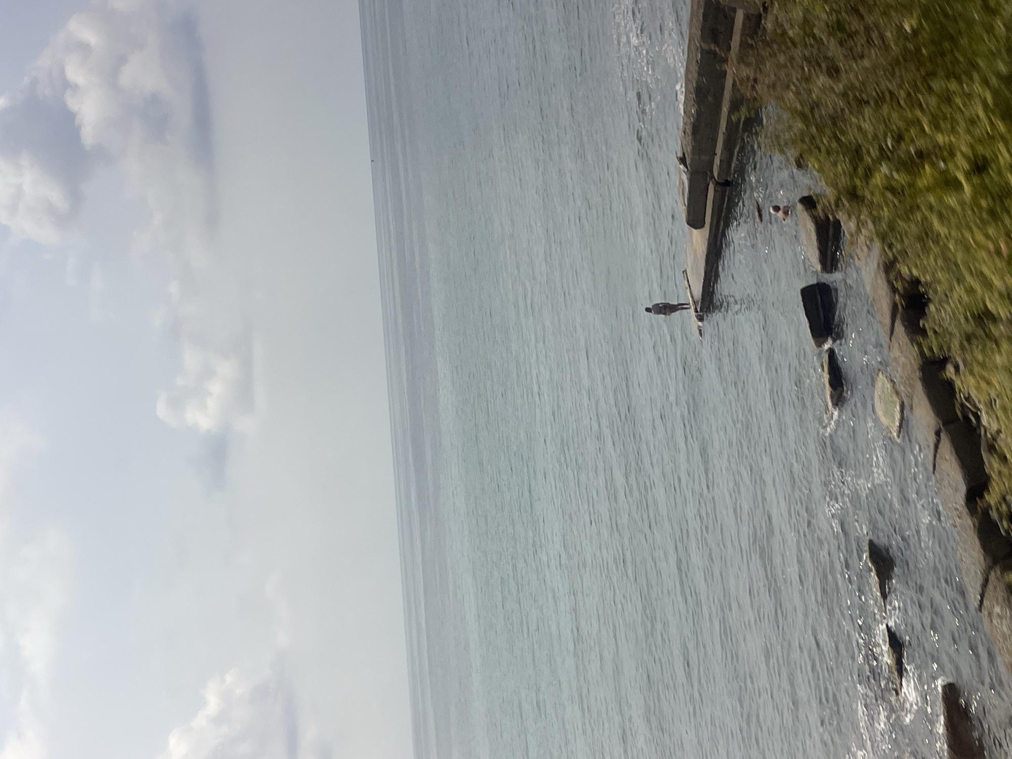 The image captures a tranquil scene at the Black Sea coast in Sochi, Russia. The expansive, calm sea stretches towards the horizon under a partly cloudy sky. In the foreground, a concrete pier juts out into the water, with a solitary figure standing on its edge, looking out at the sea. Nearby, another person is partially submerged in the water, seemingly swimming or cooling off. The shoreline is lined with large, grey rocks and lush green vegetation, adding a natural element to the urban coastal landscape. The overall impression is one of peaceful recreation and the serene beauty of a summer day.