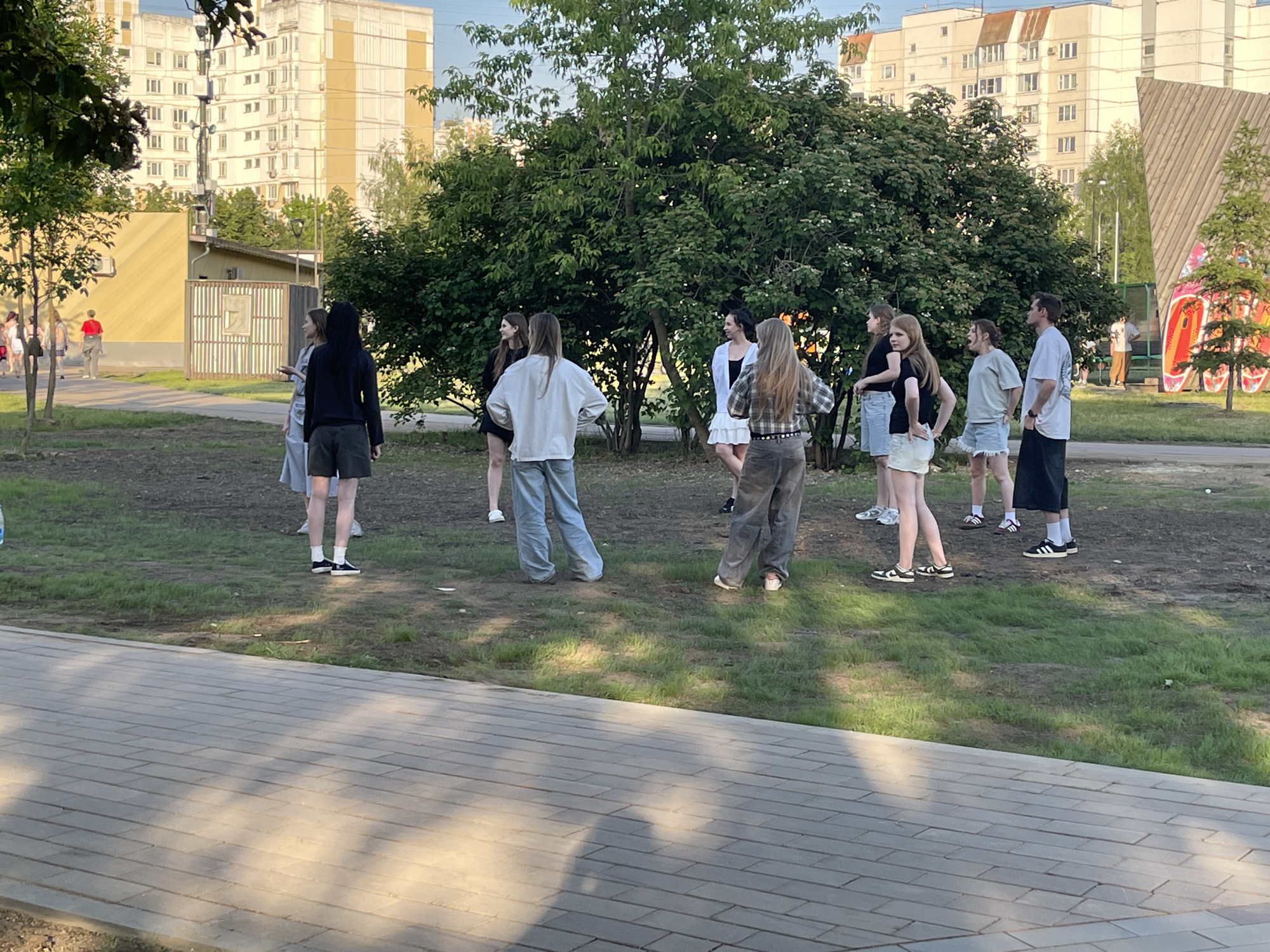 This image depicts an outdoor gathering of approximately 14 young adults in a park-like area on a sunny late afternoon or early evening. The location is identified as Pryamikov Square in Moscow, Russia, evidenced by the distinctive colorful graffiti structure visible in the background on the right. In the foreground, a paved pathway made of light-colored rectangular pavers stretches across the bottom of the frame, partially covered by long shadows from unseen objects to the left. The midground consists of a grassy area with some worn, bare patches of earth, where the majority of the individuals are standing. Several mature trees and dense green bushes are clustered in the center and right midground, casting additional shadows. Twelve to fourteen young adults, predominantly female, are distributed in two loose groups. One group of approximately five individuals is located in the left-center. One woman in a black top and dark shorts, wearing white socks and black sneakers, faces away from the viewer. Another woman in a light top and light-wash jeans also faces away. In the far left background, several more individuals, including one wearing a red shirt, are visible walking on a path. The second, larger group of approximately seven individuals is situated in the right-center. This group includes a woman in a white top and short white skirt, a woman in a plaid shirt and dark pants, and a woman in a black t-shirt and light blue denim shorts, all facing left. A woman in a light green t-shirt and light denim shorts stands with her mouth open, appearing to speak animatedly, facing towards the right. Nearby, a man in a white t-shirt and dark pants stands facing left, and another woman in a black t-shirt and light blue shorts stands facing left. A man in a dark t-shirt and dark pants, wearing white socks and black sneakers, is positioned towards the far right of this group. The individuals appear to be engaged in conversation. The background features several large, multi-story residential apartment buildings, predominantly light yellow/beige and white, with numerous windows, typical of Soviet-era architecture. A cellular tower or communication mast is visible between two of these buildings in the far left-center. To the right, a prominent, sloped wooden structure covered in large, colorful graffiti (featuring red, yellow, blue, and black outlines) stands vertically, confirming the location as a youth area within Pryamikov Square. A man in a light
