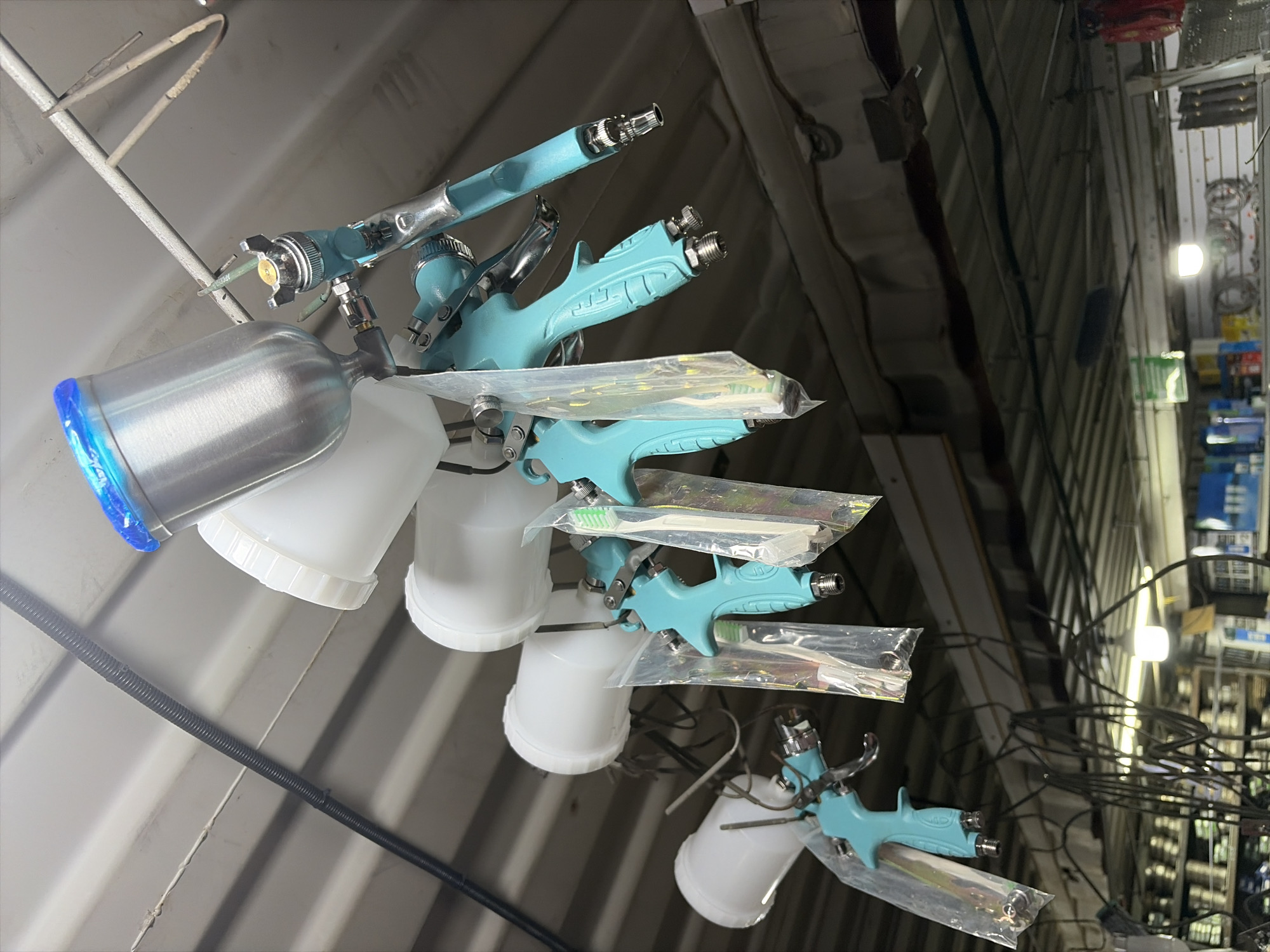 A close-up, low-angle shot captures a series of turquoise spray guns with white containers hanging from a ceiling in what appears to be a workshop or retail environment. The spray guns are suspended by wires and hooks, with some of them individually bagged in clear plastic. The topmost spray gun features a bright blue lid on its container. In the background, shelves stocked with various items and fluorescent lighting are visible, suggesting a commercial setting. The overall impression is one of organized inventory or display. The location in Kant, Kyrgyzstan is not discernible from the image itself.