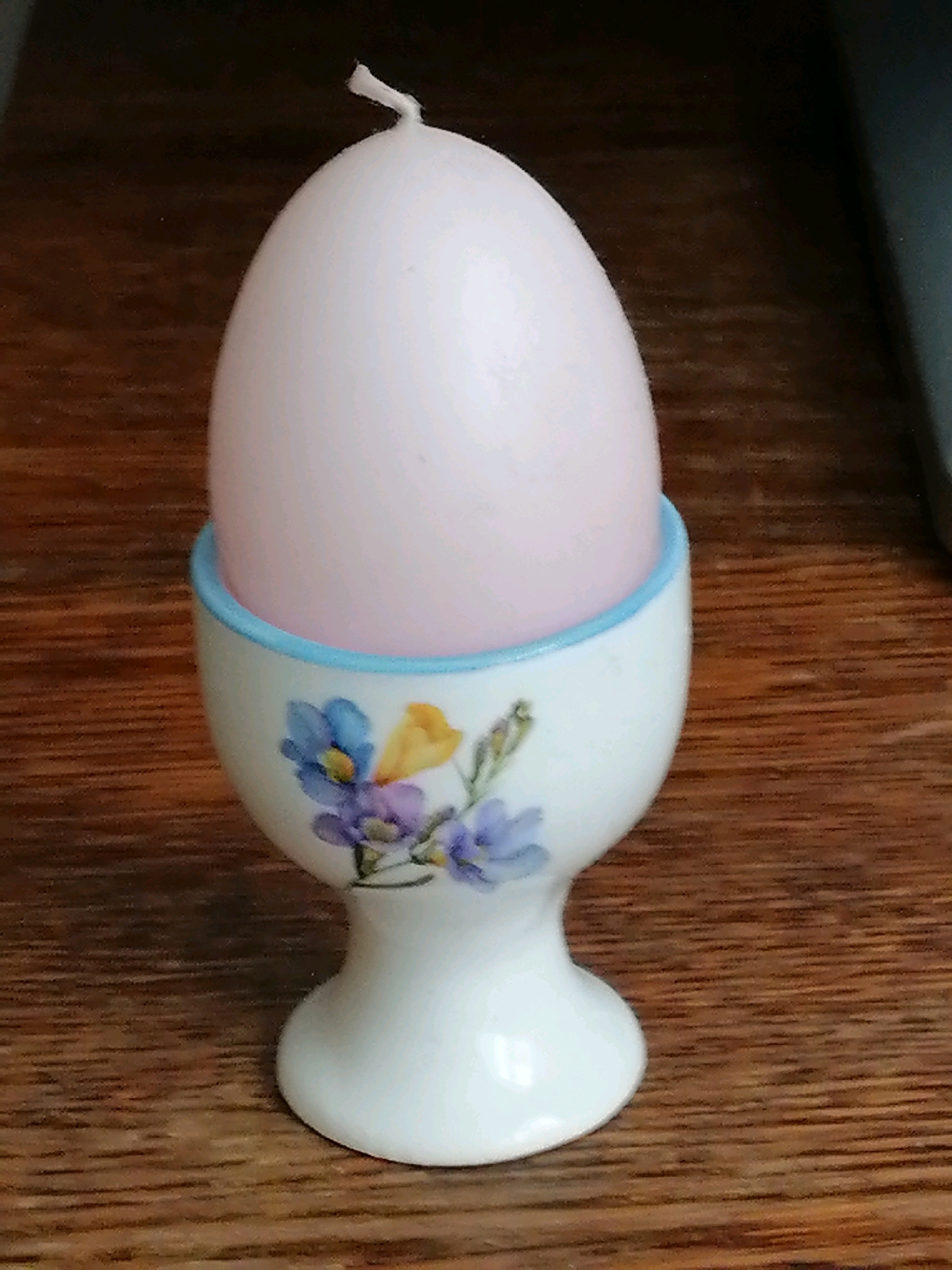 A pale pink egg-shaped candle stands in a white ceramic egg cup decorated with blue, purple, and yellow flowers. The egg cup has a light blue rim. The candle has a short white wick extending from its top. The egg cup and candle rest on a wooden surface with a visible grain pattern. The background is dark and out of focus, with a hint of a dark blue object to the right. The image is taken from a slightly high angle. There are no people or animals present.