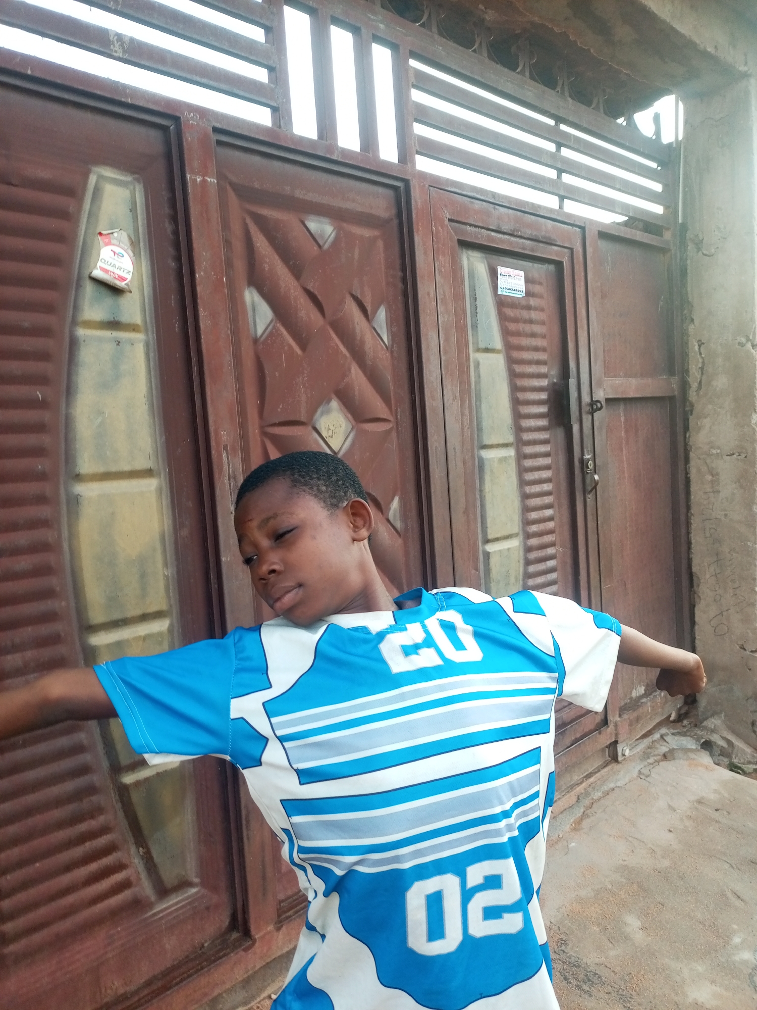 A young boy, appearing to be of African descent, stands in front of a brown metal gate in Tempure, Nigeria. He is wearing a blue and white t-shirt with the numbers "20" and "02" visible on it. His arms are outstretched, and he is looking to his left with his eyes partially closed. The gate is composed of two panels with decorative diamond patterns and horizontal slats above. A sticker is attached to the left panel of the gate. The ground in front of the gate is concrete and appears worn. The overall lighting suggests an outdoor, daytime scene.