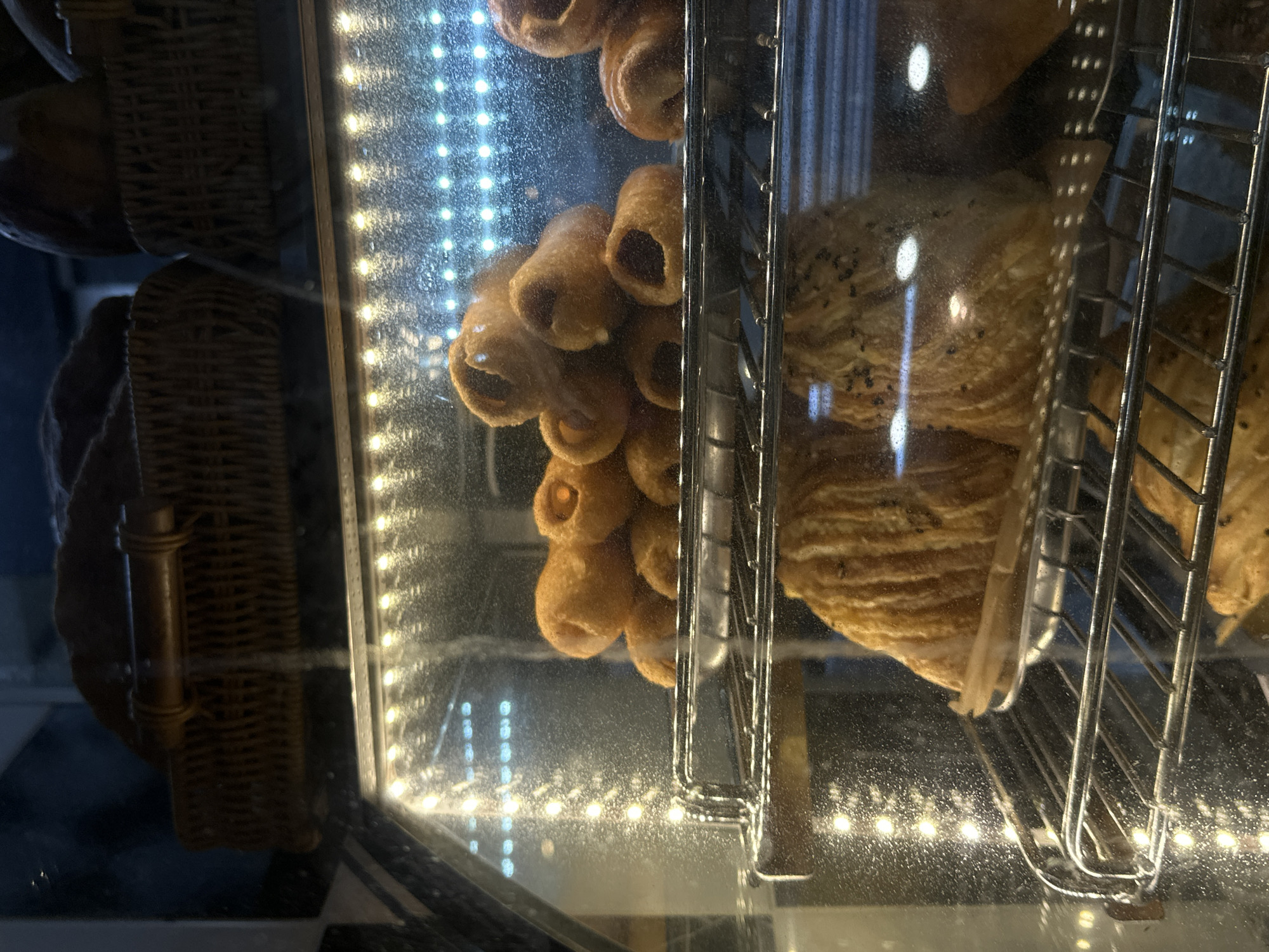 Inside a bakery display case, presumably in Moscow, Russia, two varieties of pastries are visible. On the left, a cluster of tubular pastries are arranged vertically, their dark fillings visible at each opening. To the right, layered pastries, possibly croissants, are displayed on a metal rack. The display case is illuminated with rows of small lights along the edges. Wicker baskets with additional baked goods are visible, though blurry, in the upper left and lower left corners.