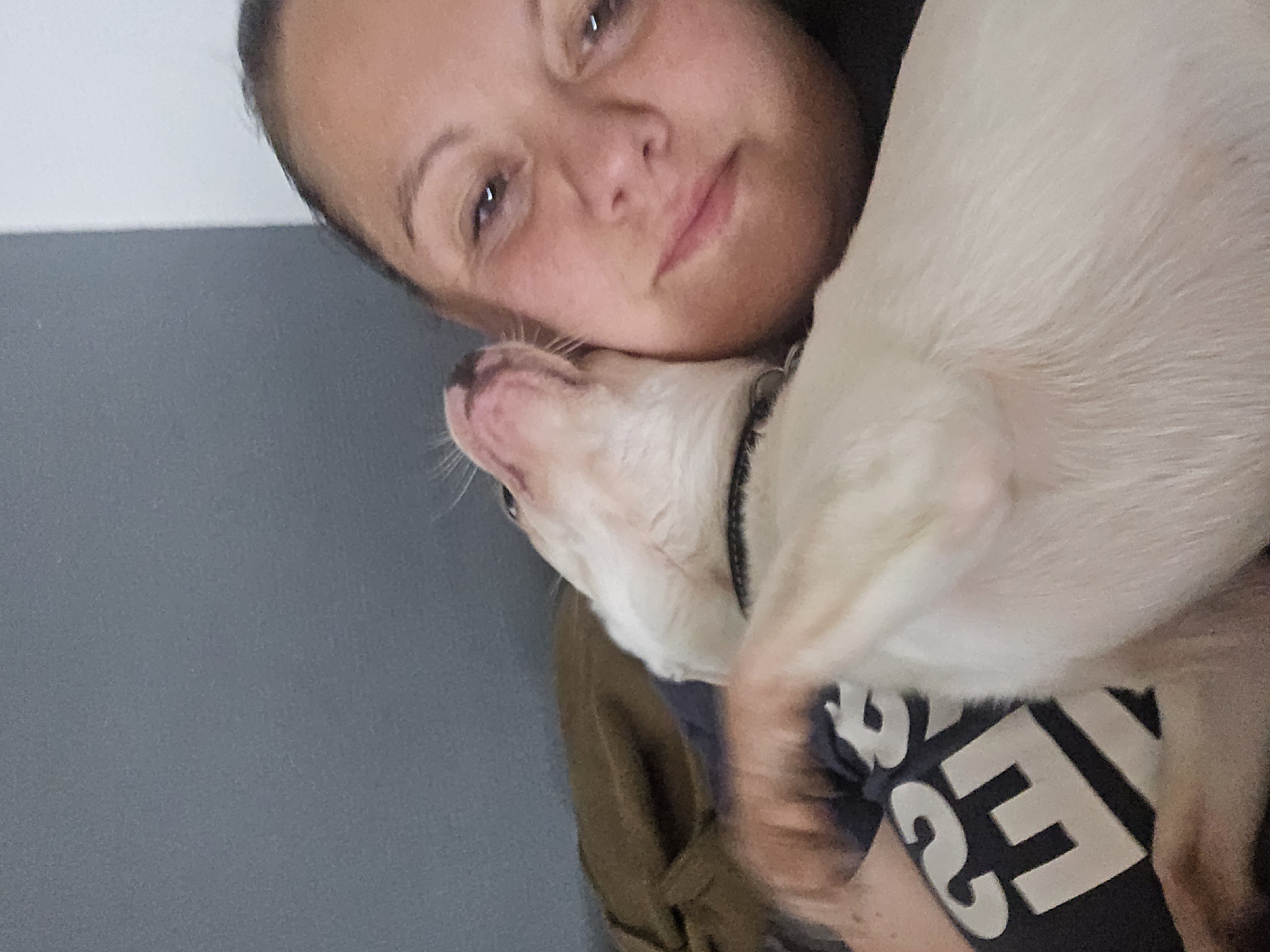 A selfie shows a woman holding a light-colored dog. The woman has fair skin, brown eyes, and dark hair pulled back. She is wearing a dark t-shirt with white lettering. The dog is pressed against her face and chest. The background is a plain wall, with a gray lower portion and a white upper portion. The location in the city of Hem, France, cannot be determined from the image.