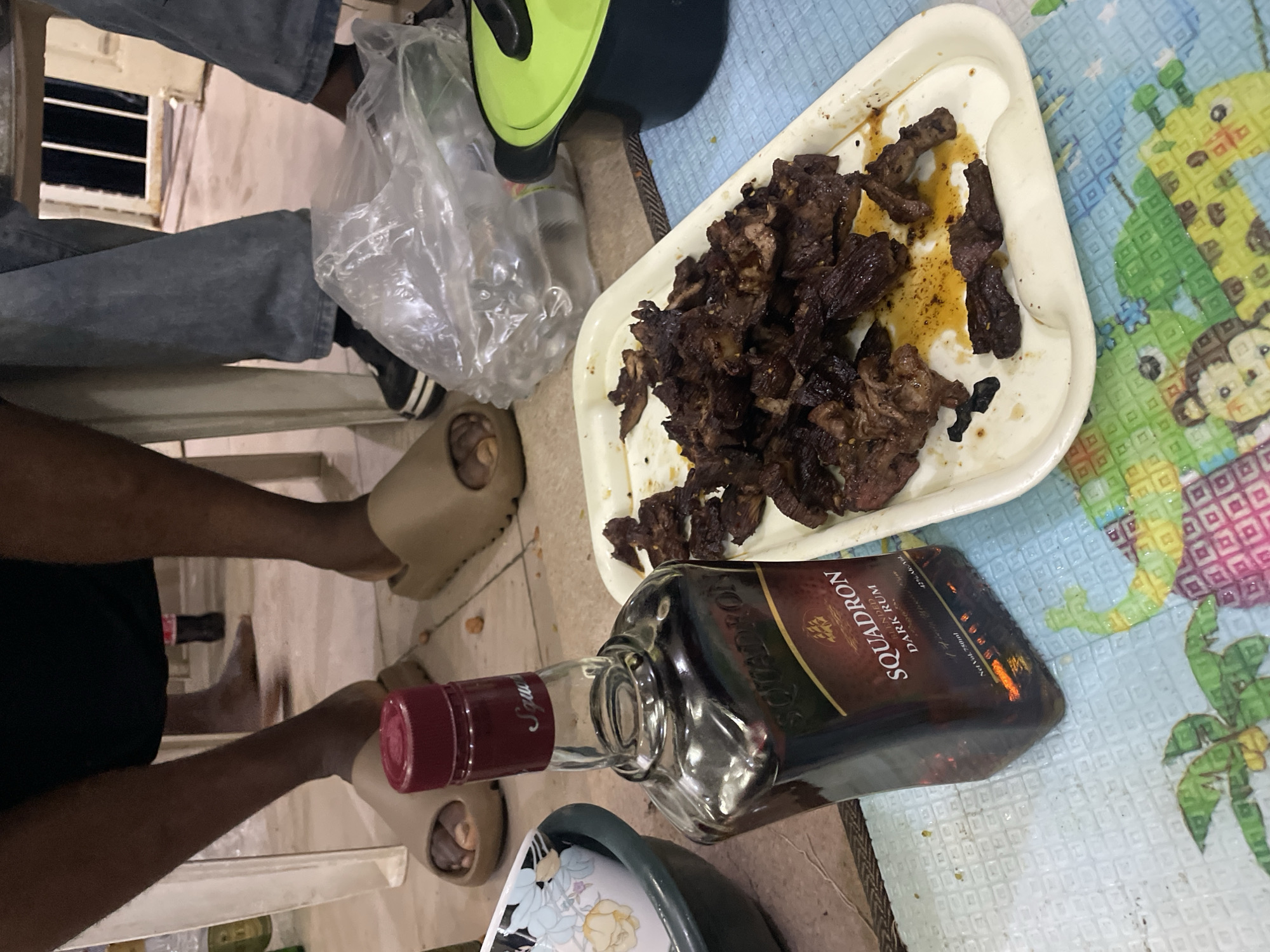 The image depicts a casual indoor scene, possibly in a home in Jos, Nigeria, given the presence of Squadron Dark Rum, a locally popular spirit. There are two people visible, one wearing jeans and sneakers, and another wearing a black shirt and tan slides. A white plastic tray holds pieces of dark, cooked meat. A partially empty bottle of Squadron Dark Rum lies horizontally on the table. Nearby are a green and black object, a plastic bag, and a floral plate. The scene is set on a table covered with a patterned tablecloth.