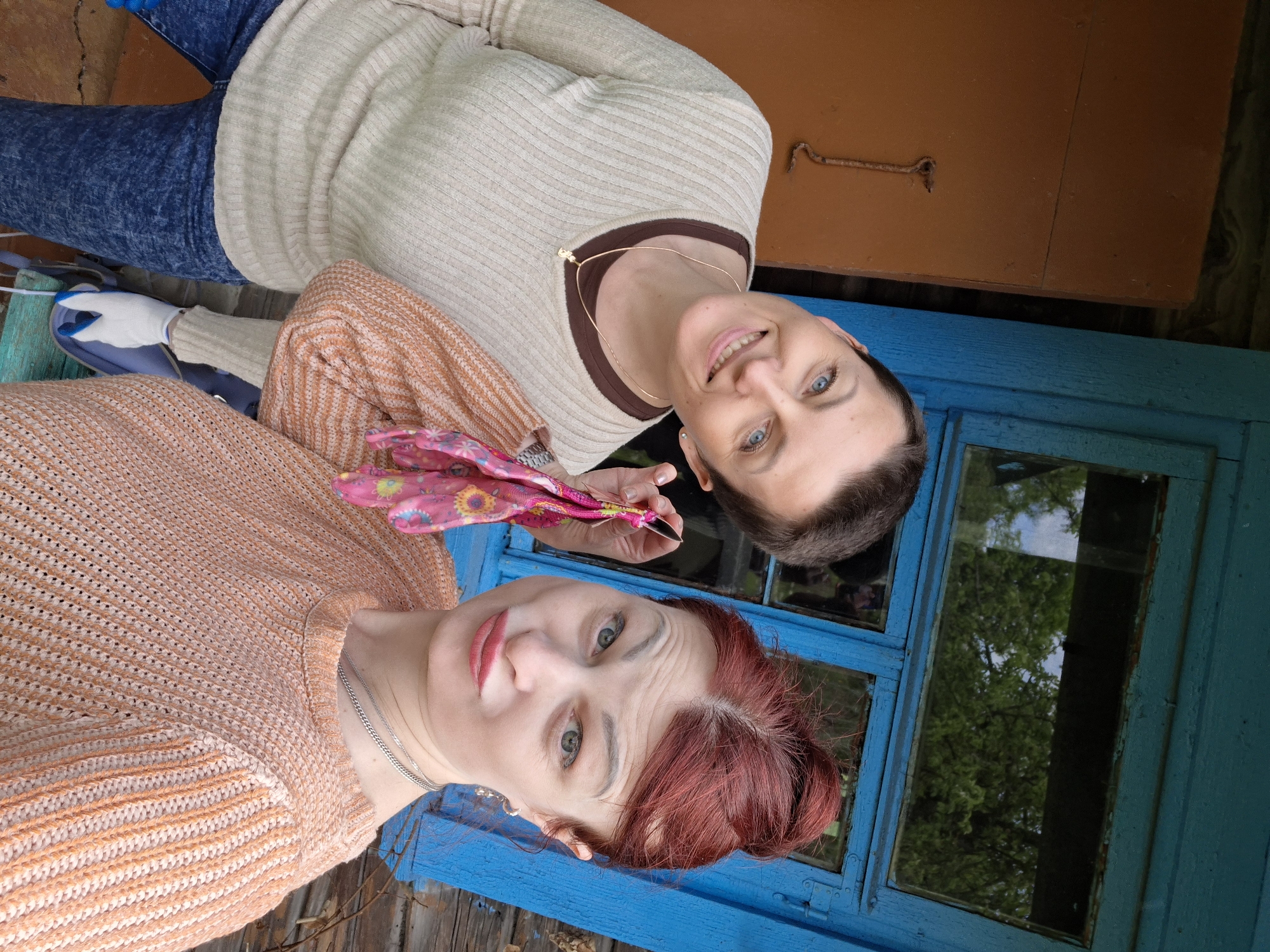 Two women are posing for a photo. The woman on the right is wearing a beige sweater with brown trim and a gold necklace. The woman on the left is wearing an orange sweater and is holding a pair of pink gardening gloves. They are standing next to a blue window and door. The location is likely Сваткаўскі сельскі Савет, Belarus based on the user's location.