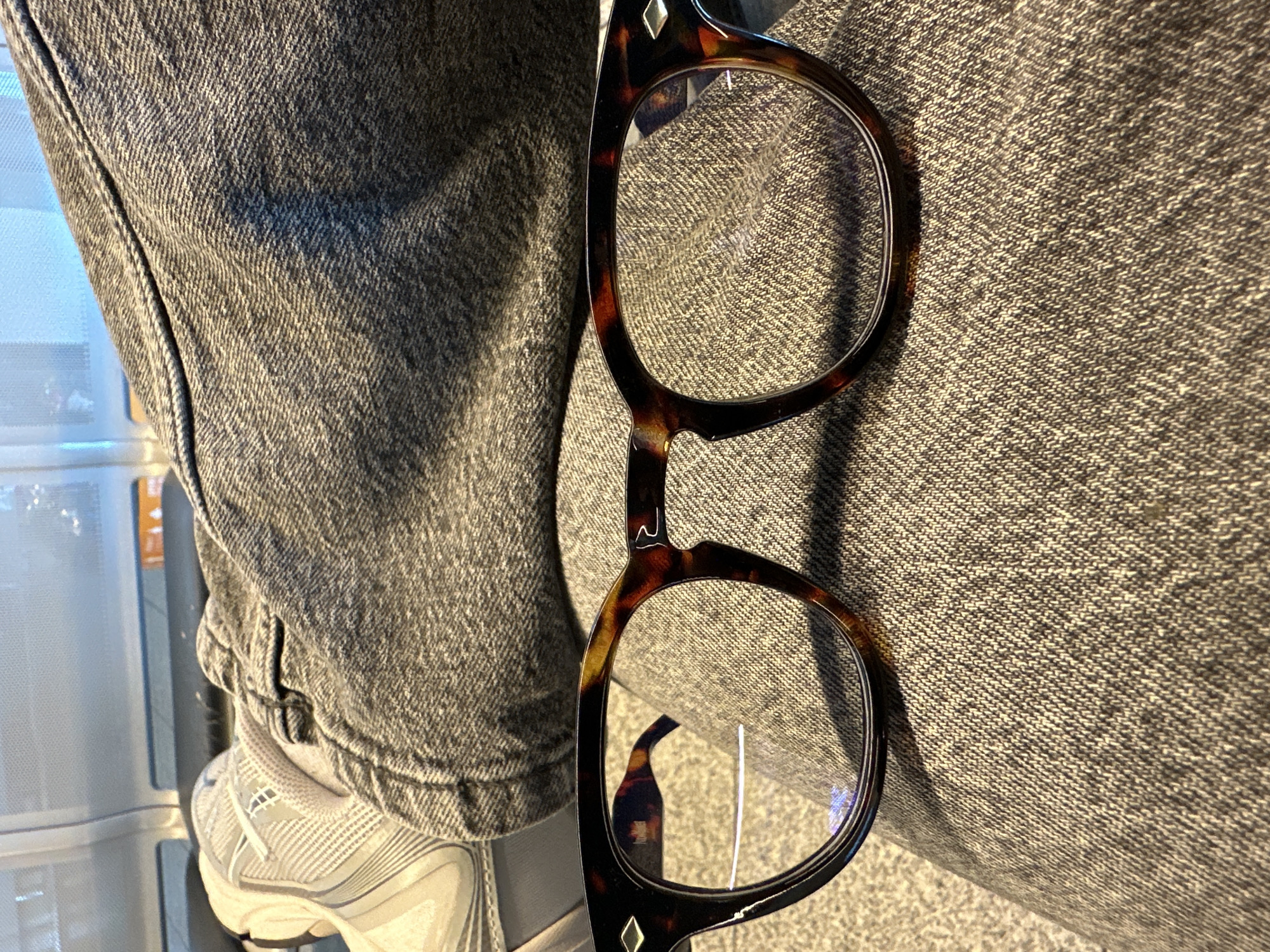 This close-up image, captured in Weeze, Germany, features a person's leg clad in dark grey denim jeans and a white athletic shoe, with a pair of tortoiseshell eyeglasses resting prominently on the fabric. The denim displays a distinct twill weave texture with subtle folds. On the lower left, a white sneaker with grey stripes and accents is visible, though its brand logo is indistinct due to blurriness.
The eyeglasses, the central subject, boast stylish dark brown and amber mottled frames. A small, silver-colored diamond-shaped inlay decorates the upper corner of the right lens frame. On the bridge connecting the two lenses, a unique, stylized symbol resembling a wave or an 'M' is etched. The clear lenses show a slight blue reflection, possibly indicating blue light filtering or an anti-reflective coating.
The background is softly blurred, revealing light-colored, linear structures, possibly shelving or a part of a cabinet, suggesting an indoor setting. The image captures a moment of stillness, with the items appearing to be temporarily placed. No specific activity is taking place, nor are any emotions discernible. The lighting appears to be ambient indoor light, but the time of day is not evident. No explicit text is clearly legible, apart from the stylized symbol on the eyeglasses.