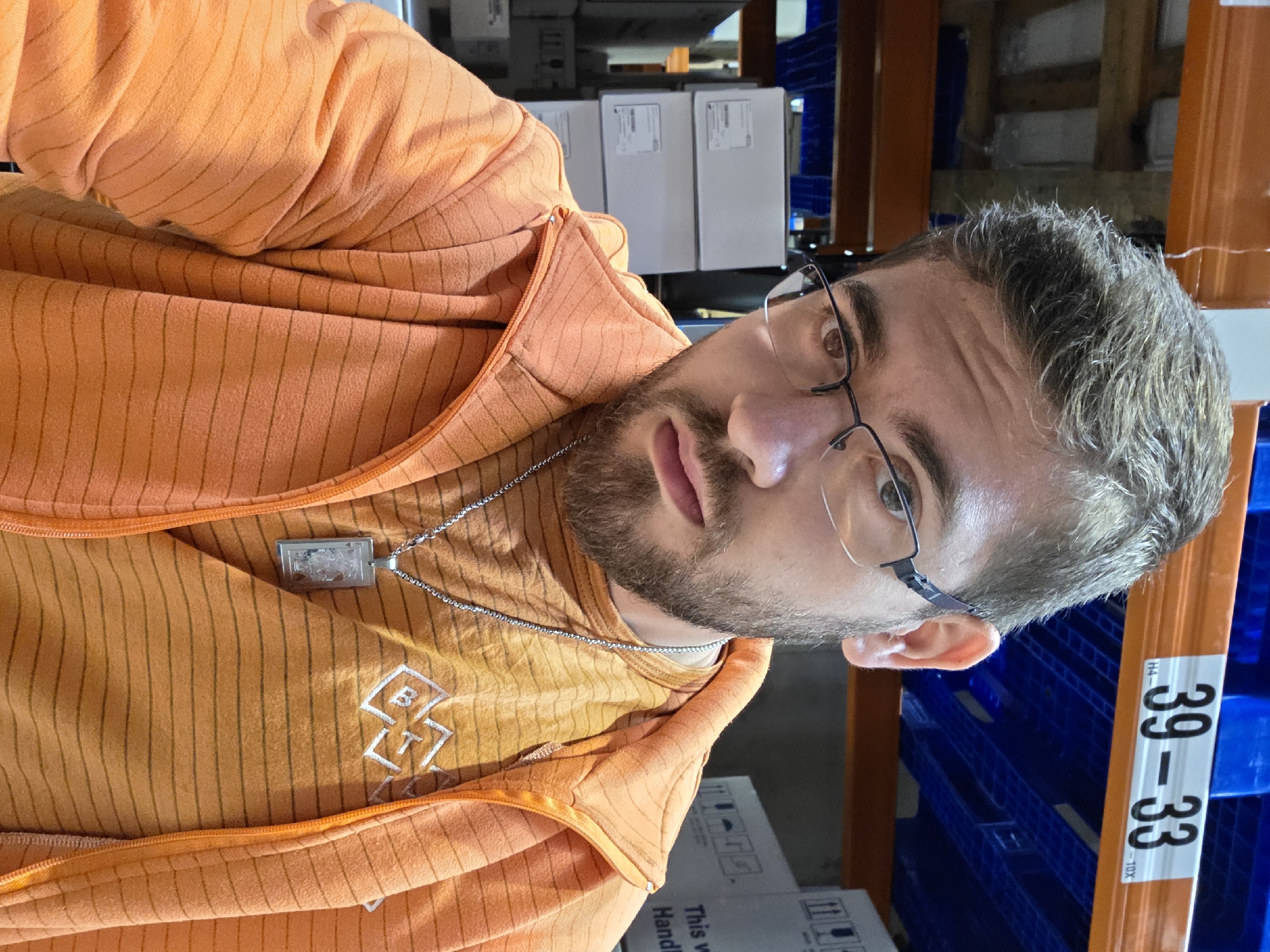 A man is standing in a warehouse. He is wearing an orange striped shirt and jacket, glasses, and a necklace with a pendant. Boxes are on shelves behind him. The warehouse appears to be in Plovdiv, Bulgaria based on the labels on boxes in the background.