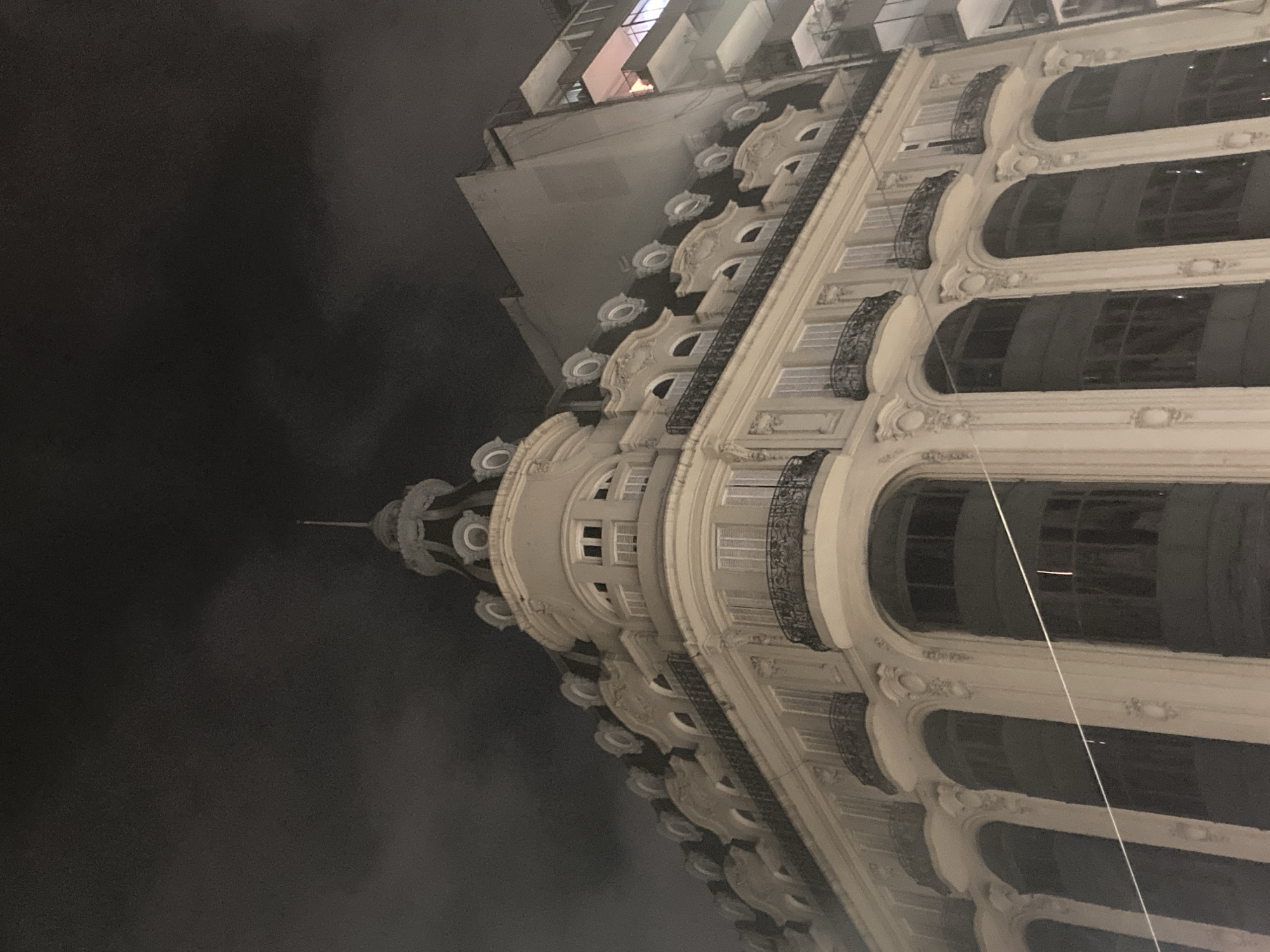 The image captures a large, ornate building at night, viewed from a low angle looking upwards. The building displays classical architectural elements with a light-colored, possibly stone or stucco, facade. Intricate detailing is visible around the numerous arched windows, which appear dark with some internal reflections or faint lights. Many windows are accompanied by decorative balconies featuring dark, wrought-iron railings.
The building's prominent feature is a rounded corner that rises to a multi-tiered dome or turret, topped with a distinct spire or finial. The structure is illuminated, highlighting its elaborate cornices, columns, and other decorative flourishes against a dark, cloudy, or hazy night sky. In the background, to the upper left, parts of other, less detailed modern buildings are visible. A thin, light-colored line, possibly a wire or string, extends diagonally across the lower right section of the image. No people or specific activities are depicted. The time of day is clearly night, indicated by the dark sky and artificial lighting. No visible text is present.