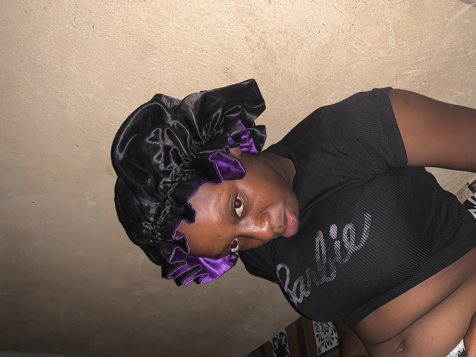 A young woman with dark skin and brown eyes wears a black satin bonnet with a purple trim. She is wearing a black ribbed t-shirt with a gray graphic design that appears to say "Believe." Her body is positioned at an angle, leaning towards the right side of the frame. The background consists of a textured, light brown wall. The lighting is dim, casting shadows on her face and creating a dramatic effect. There are no visible indications of the city or specific location.